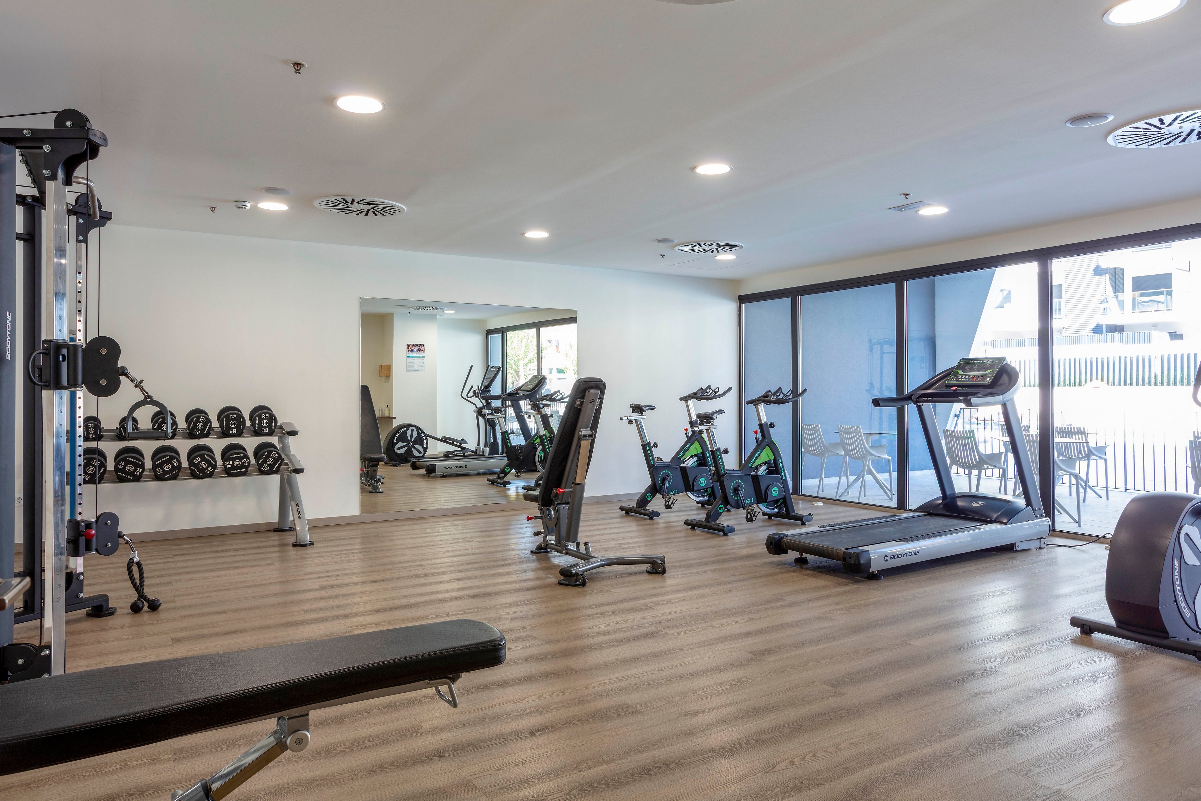 A modern fitness room features a variety of exercise equipment neatly arranged on a wooden floor. Cardio machines such as treadmills and stationary bikes are positioned near large windows that let in natural light. Free weights and dumbbells are organized on racks along the wall, with a bench and other strength-training machines in the foreground. The space appears clean, bright, and ready for individual workouts.
