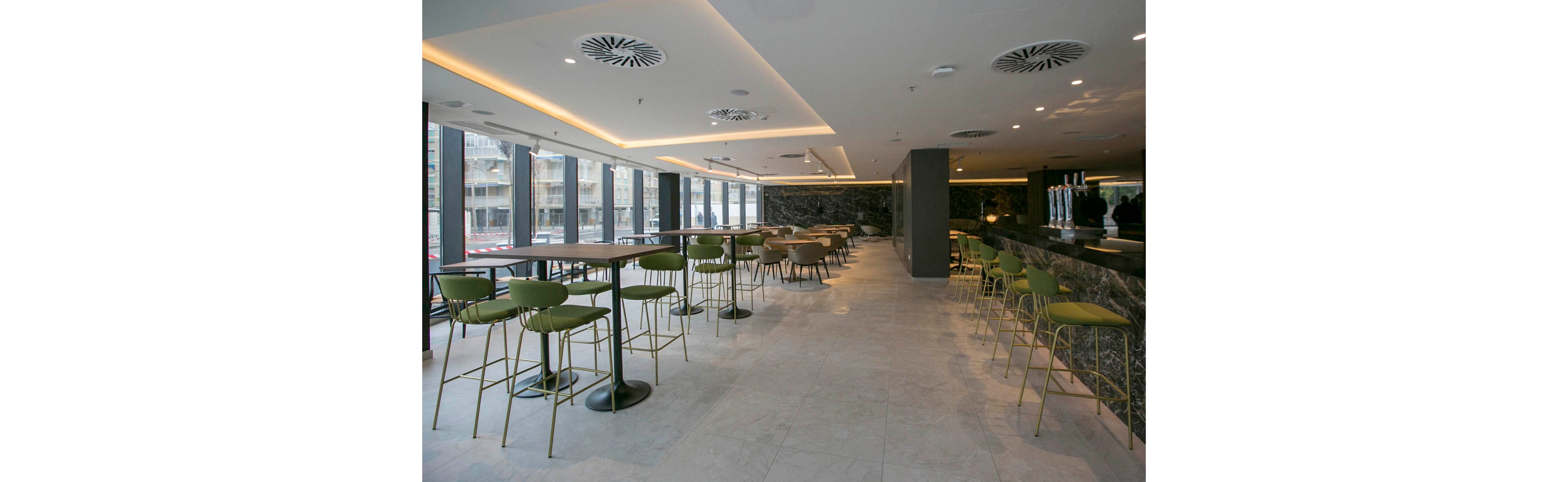 A contemporary bar lounge interior features rows of high tables and green bar stools. Large floor-to-ceiling windows line one side, allowing natural light to brighten the space and reveal an urban streetscape outside. The long bar counter runs along the opposite wall, with sleek taps and a dark stone finish. Recessed ceiling lighting and minimalist decor create a clean, modern atmosphere in the empty room.