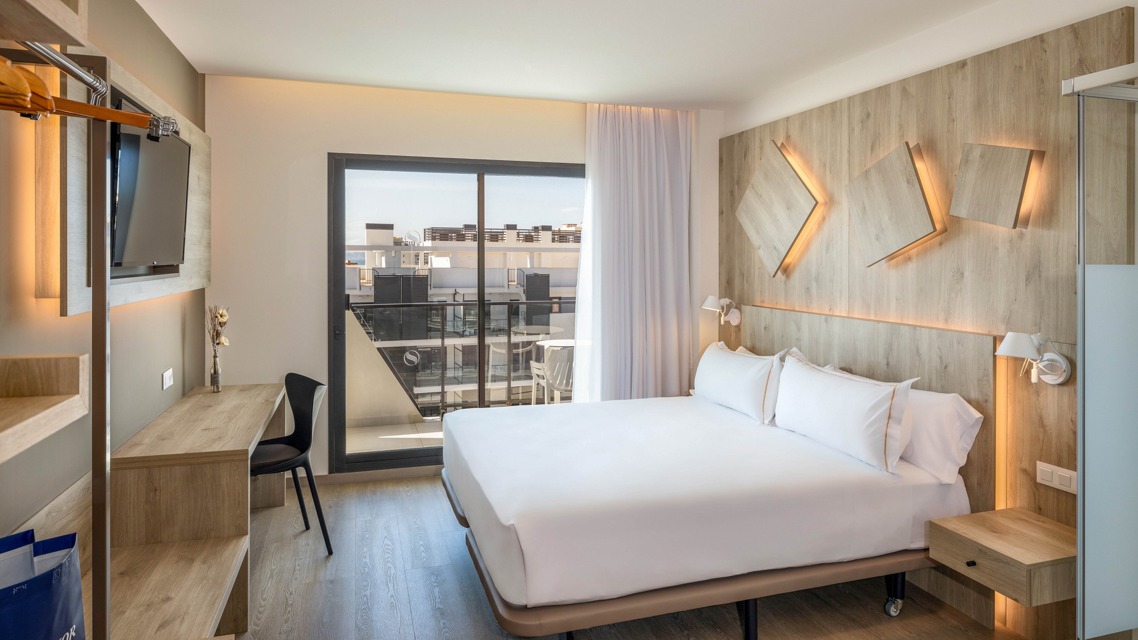 A contemporary hotel room features a large neatly made bed with white linens against a wooden accent wall. Geometric wall lights create a warm glow, complementing the natural wood furniture and flooring. A desk with a chair and wall-mounted television sit opposite the bed, providing a functional workspace. Floor-to-ceiling glass doors open to a balcony with views of neighboring buildings under a clear sky.