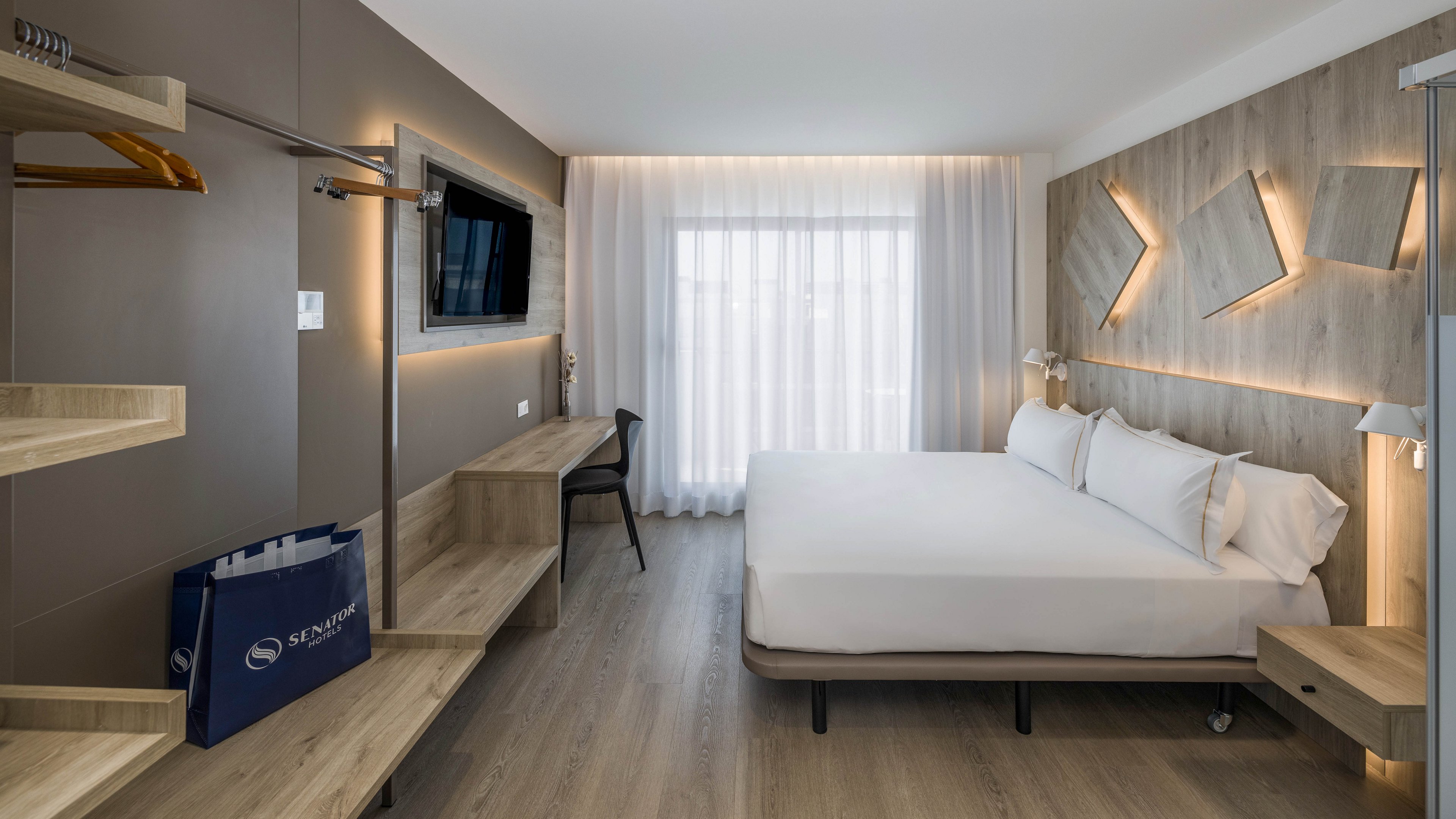 A contemporary hotel room features a large double bed with white linens against a wood-paneled accent wall. Warm integrated lighting and geometric wall fixtures create a cozy, modern atmosphere. A flat-screen television is mounted opposite the bed above a long wooden desk and bench. Floor-to-ceiling sheer curtains cover a bright window, and an open wardrobe with hangers and a blue shopping bag completes the minimalist design.