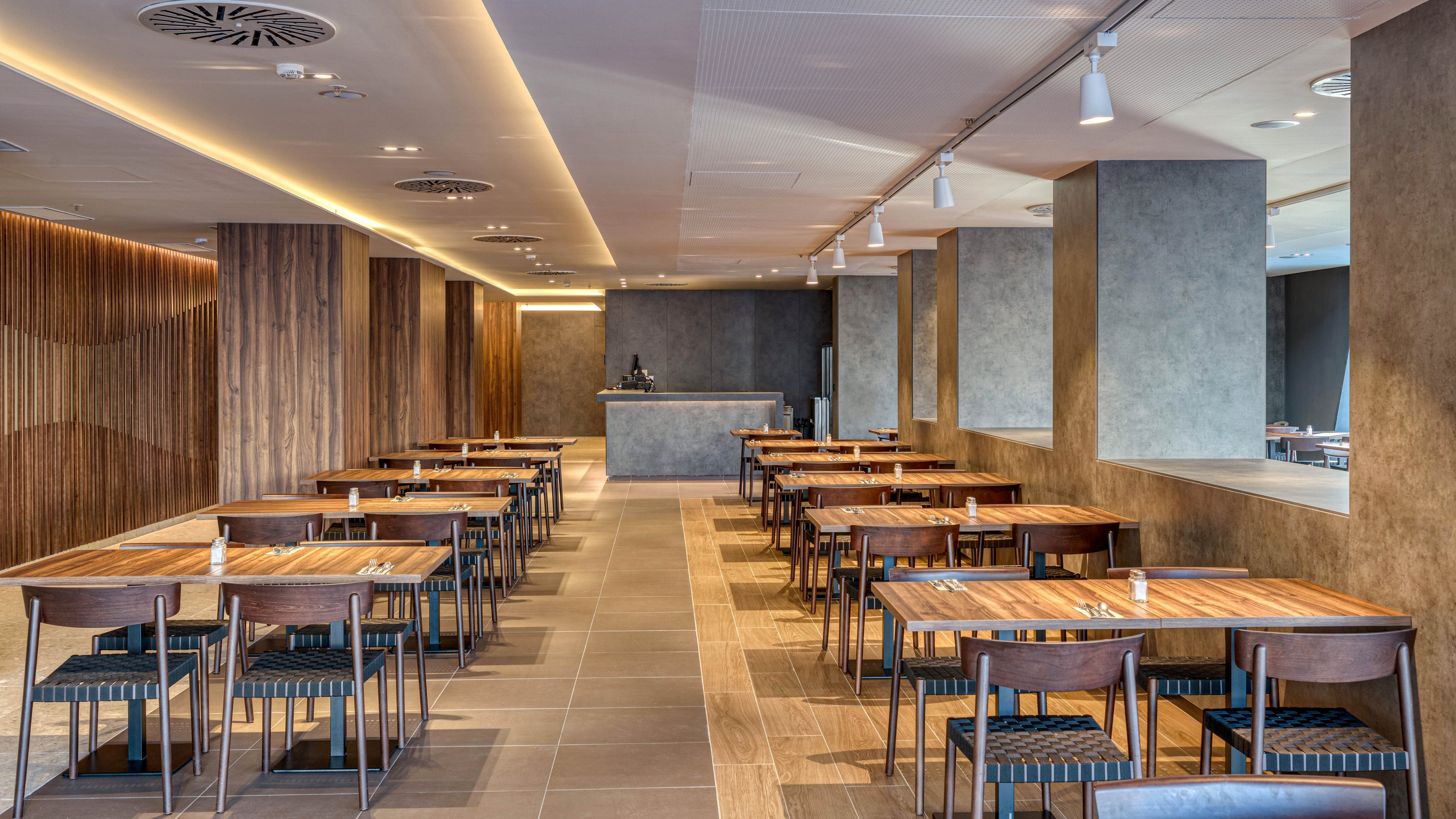 A contemporary restaurant dining area features rows of wooden tables and chairs arranged neatly in a spacious room. The setting appears empty, with no people present, emphasizing the clean and orderly layout. Warm wood paneling contrasts with smooth gray walls and columns, while recessed ceiling lights and spotlights create a soft, even illumination. At the far end, a minimalist counter or bar anchors the space, reinforcing the modern, upscale atmosphere.