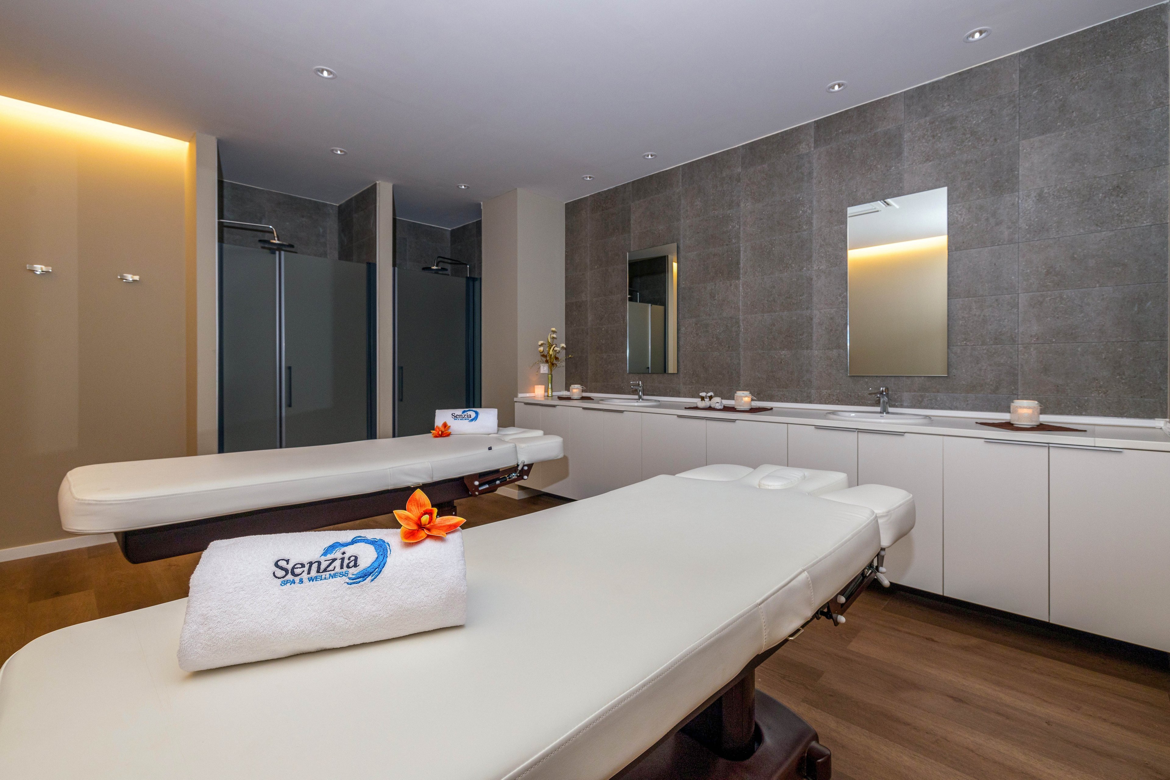A contemporary spa treatment room features two padded massage tables positioned side by side. The space includes a long white counter with sinks, mirrors, and several lit candles creating a calm ambiance. A neatly folded white towel with the word "Senzia" printed on it rests on one table, decorated with an orange flower. In the background, glass shower enclosures and warm recessed lighting enhance the relaxing, minimalist design.