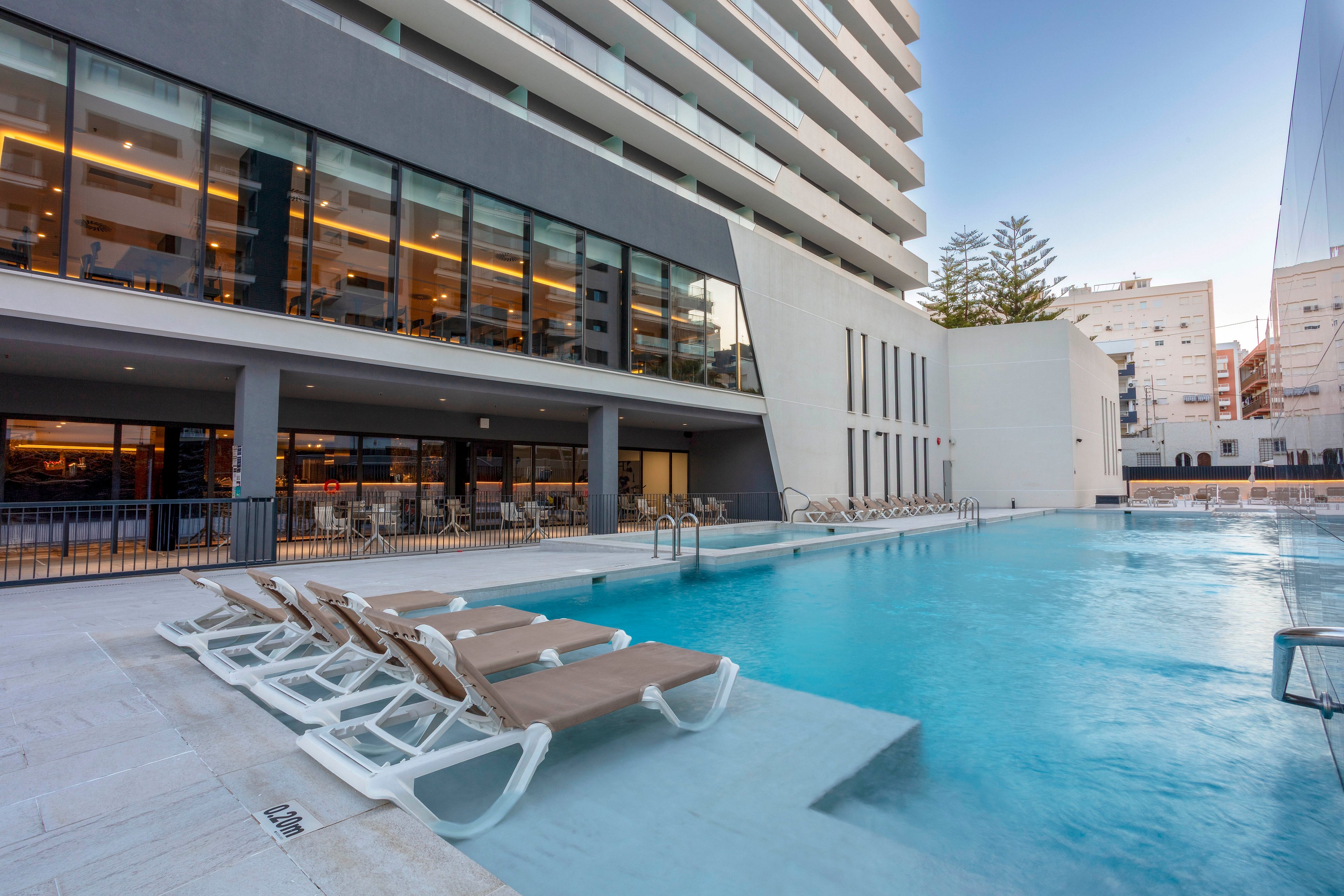 A contemporary hotel building features a large outdoor swimming pool in the foreground. Several lounge chairs are partially submerged on a shallow ledge, inviting guests to relax by the water. The hotel’s glass-fronted lower level reveals a dining or lounge area with warm interior lighting. Surrounding urban buildings and a clear evening sky frame the scene, emphasizing a calm and modern resort atmosphere.