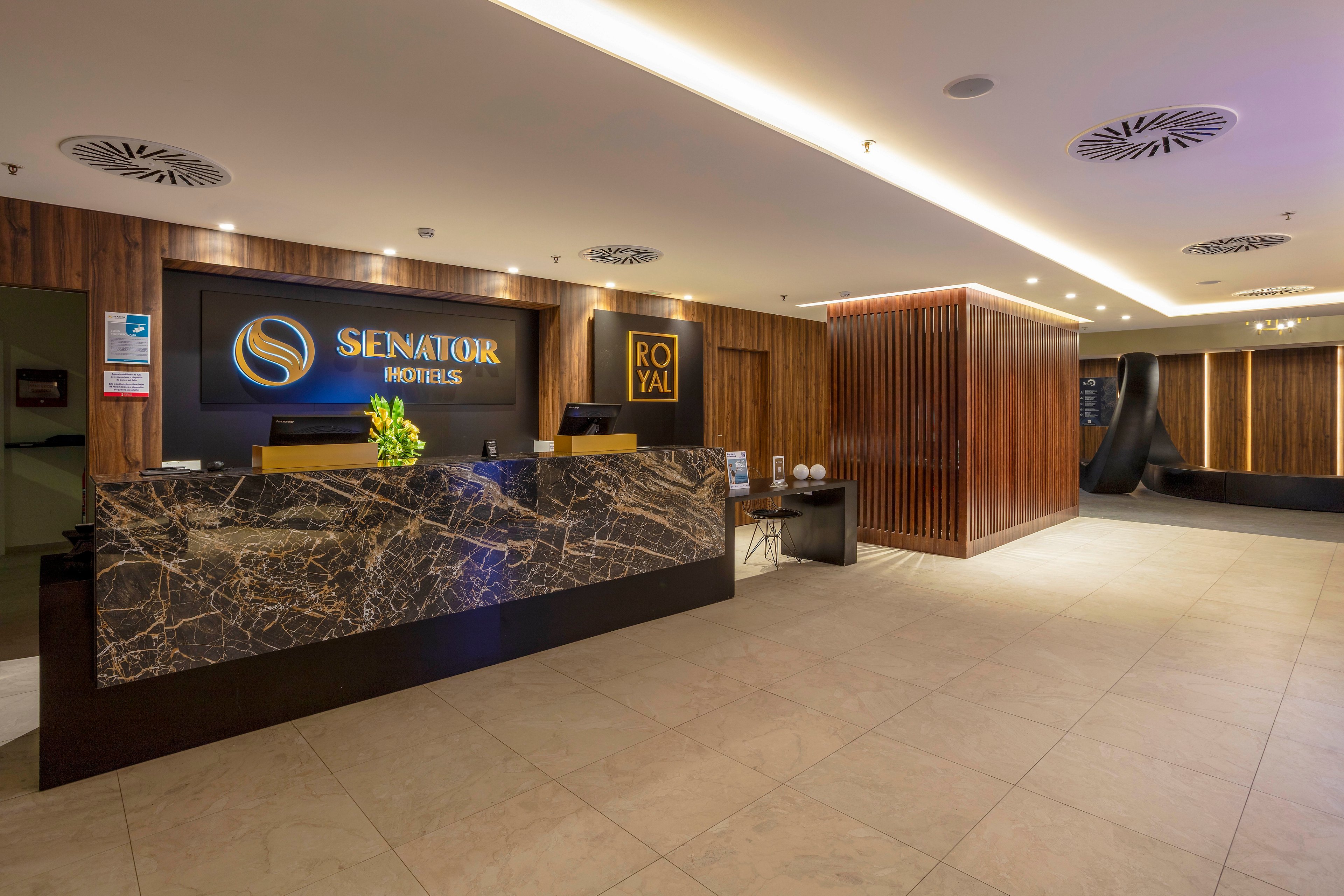 A contemporary hotel reception area features a large marble front desk with a polished dark stone surface. Warm wooden wall panels and soft ceiling lighting create an inviting atmosphere. The SENATOR HOTELS sign is prominently displayed behind the counter, alongside a ROYAL sign. Fresh flowers and informational materials sit on the counter, while the tiled floor and clean lines emphasize a modern, professional design.