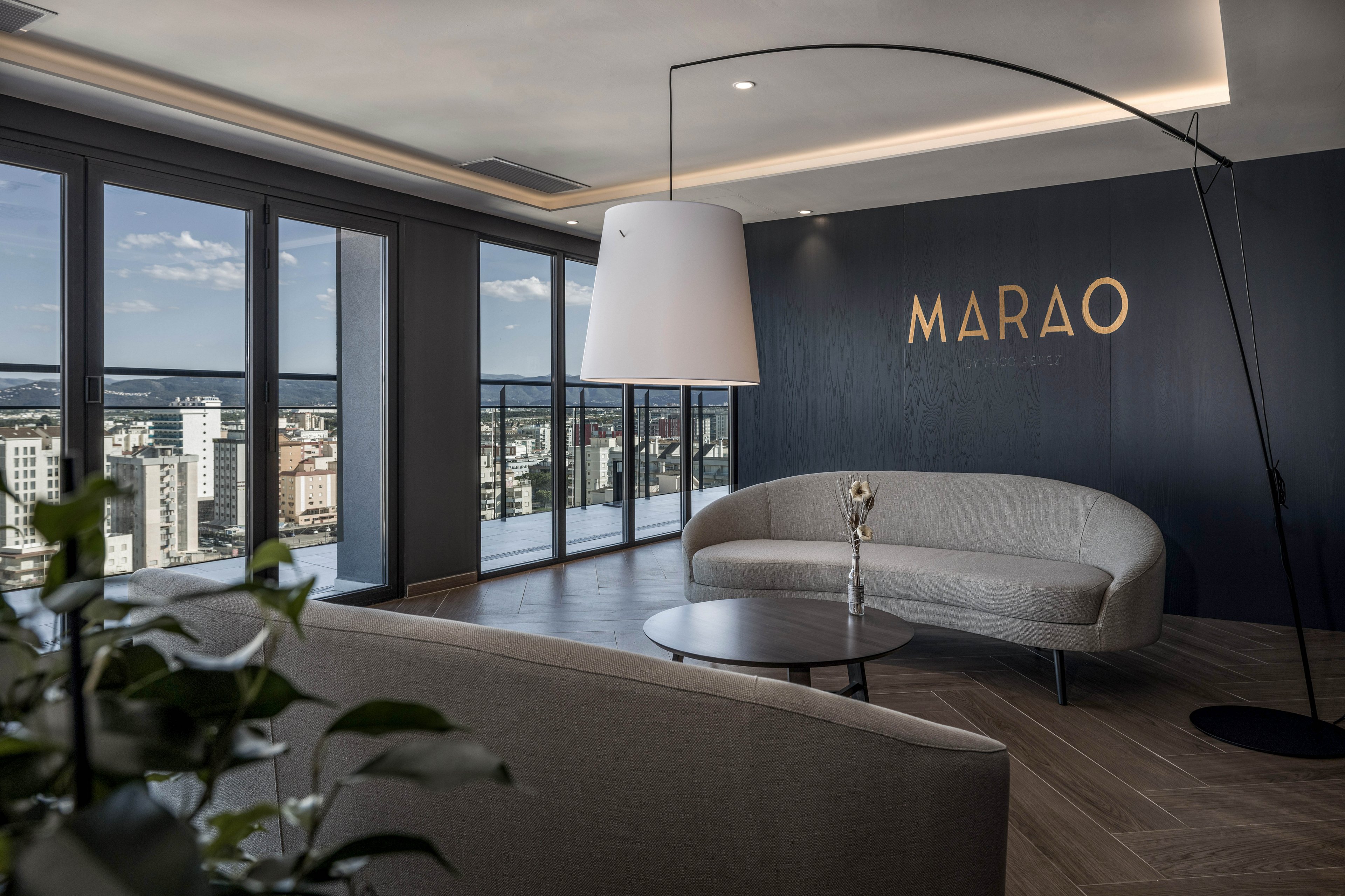 A contemporary lounge area features curved sofas and a round coffee table set against a dark accent wall. Large floor-to-ceiling windows reveal a panoramic view of a city skyline and distant horizon. The word "MARAO" is prominently displayed on the wall in warm metallic letters. Soft, indirect lighting and a minimalist floor lamp create a calm, sophisticated atmosphere. A small plant in the foreground adds a touch of greenery to the sleek interior.