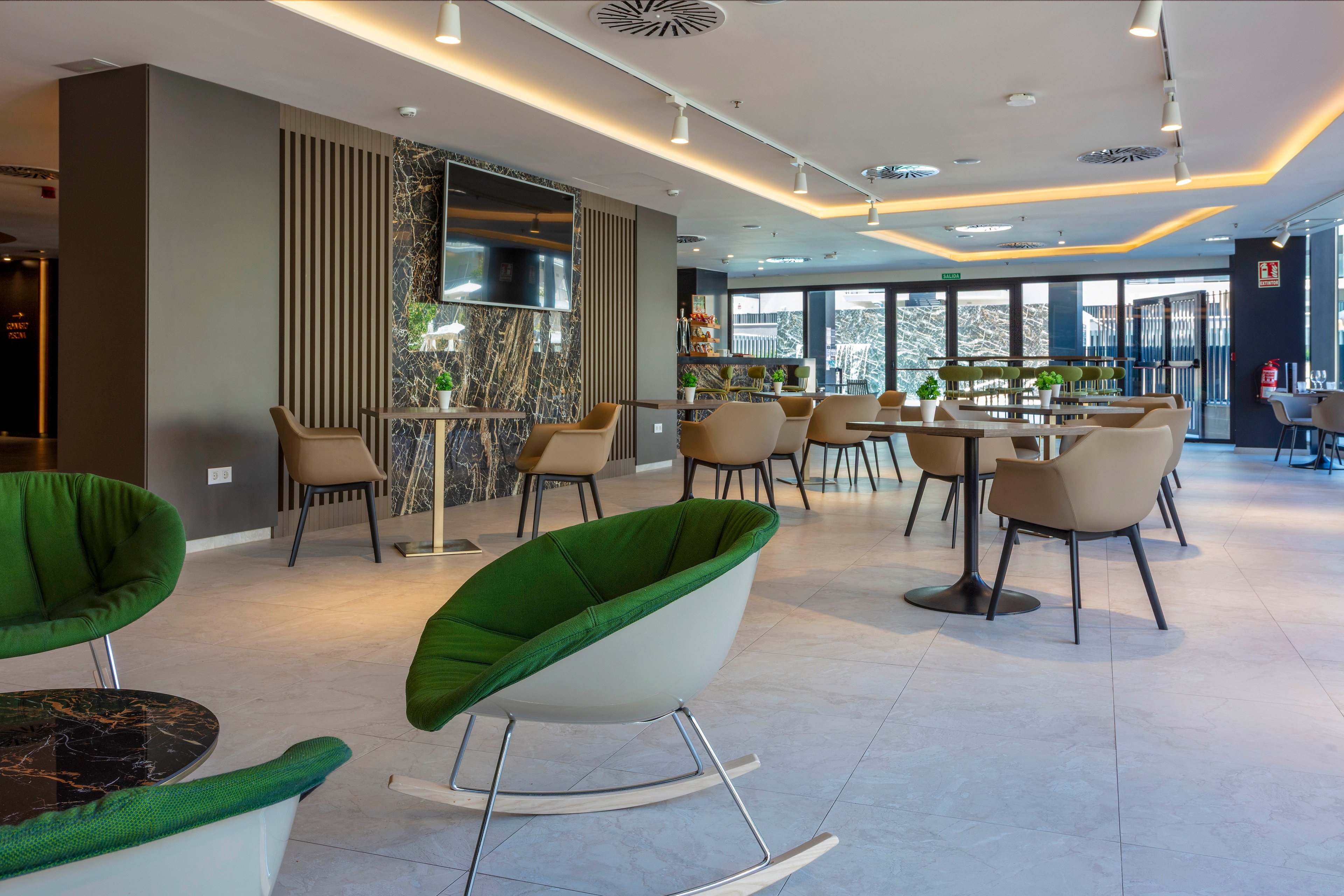 A spacious modern lounge features contemporary chairs and small round tables arranged across a bright open area. Large windows in the background provide natural light and a view of the city. The interior design includes neutral tones with green accents, marble details, and recessed ceiling lighting. The space appears clean, quiet, and suitable for casual meetings or relaxation.