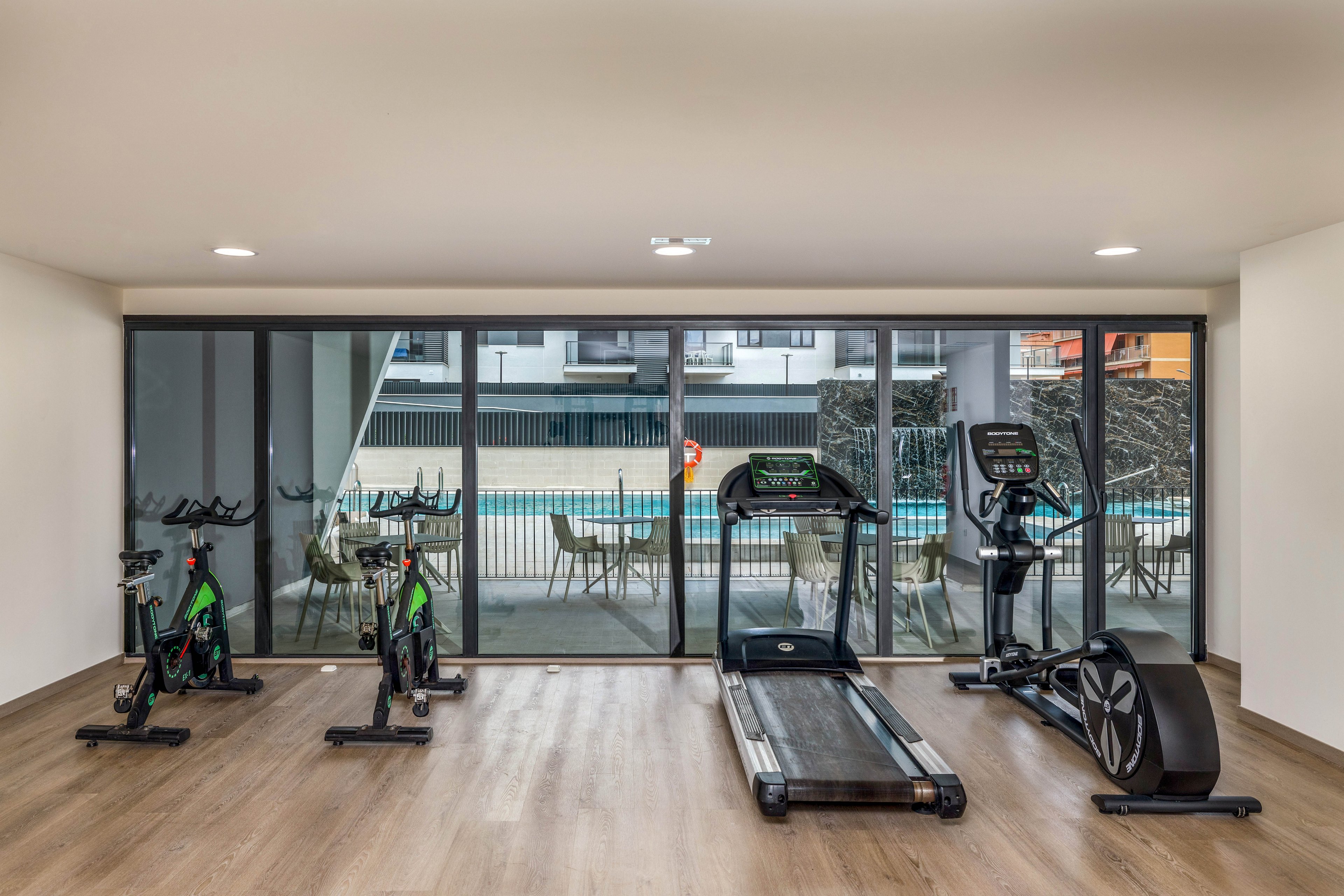 A small modern fitness room features two stationary bikes, a treadmill, and a rowing machine arranged on a wooden floor. Large floor-to-ceiling windows reveal an outdoor swimming pool area with tables and chairs. The space is bright and minimalistic, with neutral walls and recessed ceiling lights. No people are present, emphasizing the clean and orderly gym environment.