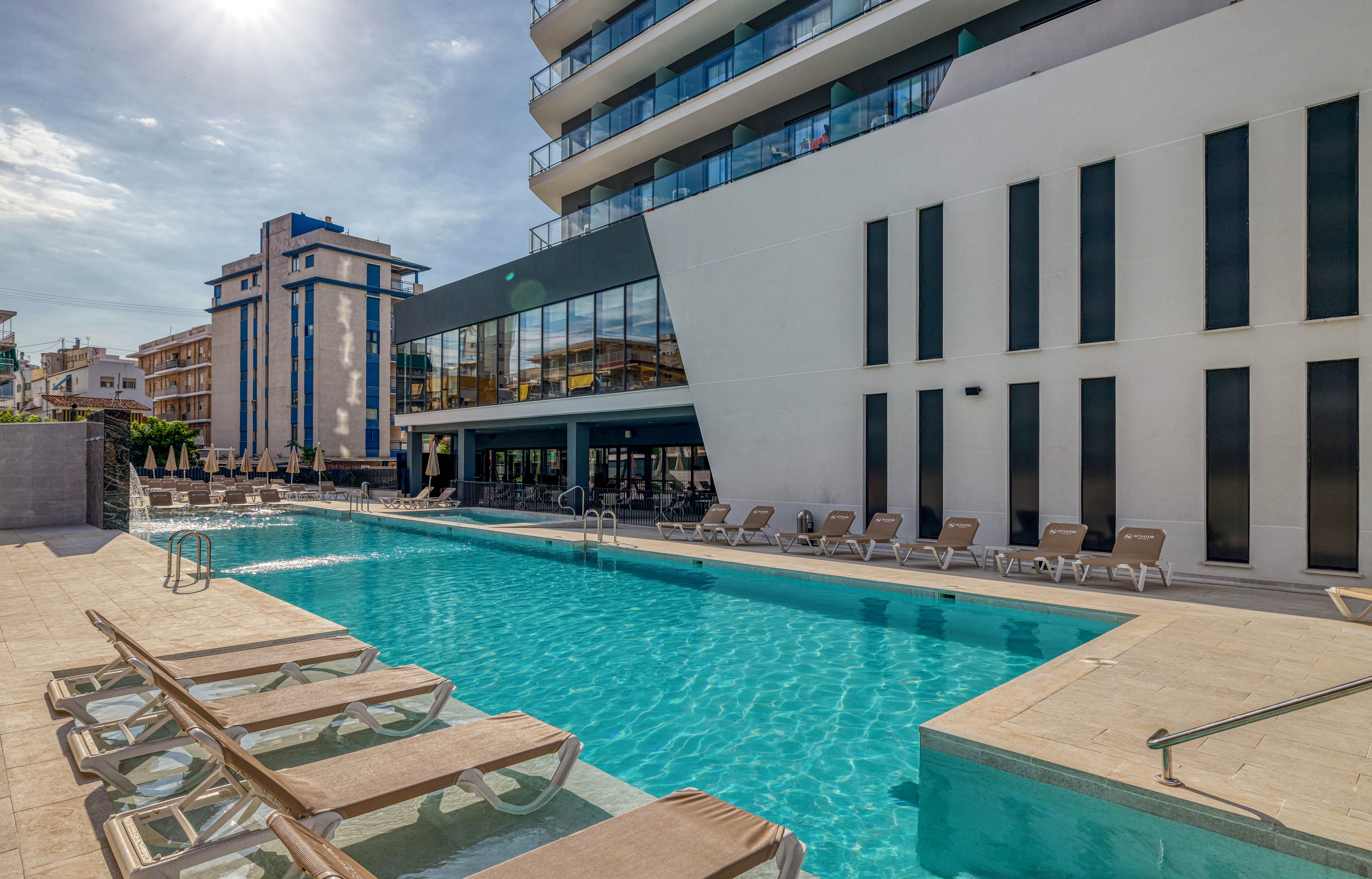 A contemporary hotel building features a long rectangular outdoor swimming pool in the foreground. Sun loungers line the poolside terrace, creating a relaxed resort atmosphere. The architecture is sleek and modern, with large glass windows and vertical black accents on a white facade. Nearby urban buildings and a clear sky suggest a sunny city or coastal destination.