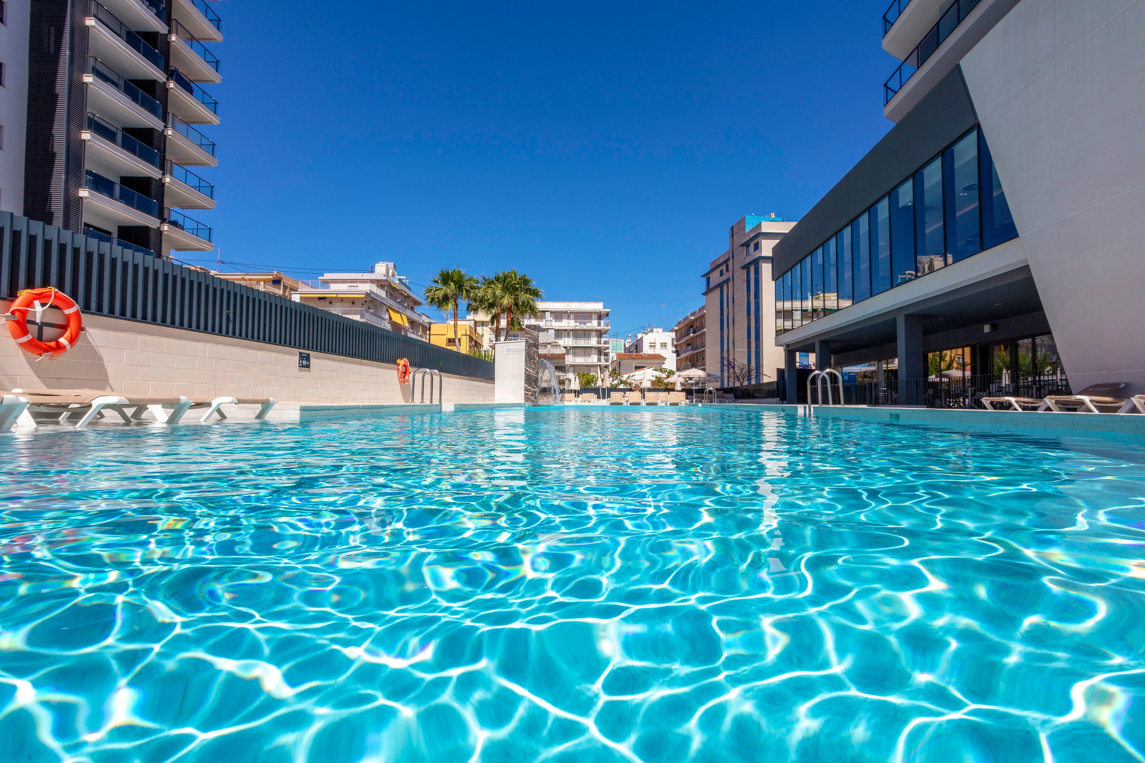 A bright outdoor swimming pool stretches across the foreground of a modern hotel complex. Clear turquoise water reflects sunlight, creating shimmering patterns on the pool surface. Contemporary buildings and balconies frame the scene, with a few palm trees visible in the background. A life preserver and pool ladders emphasize the recreational and safety features of this sunny resort setting.