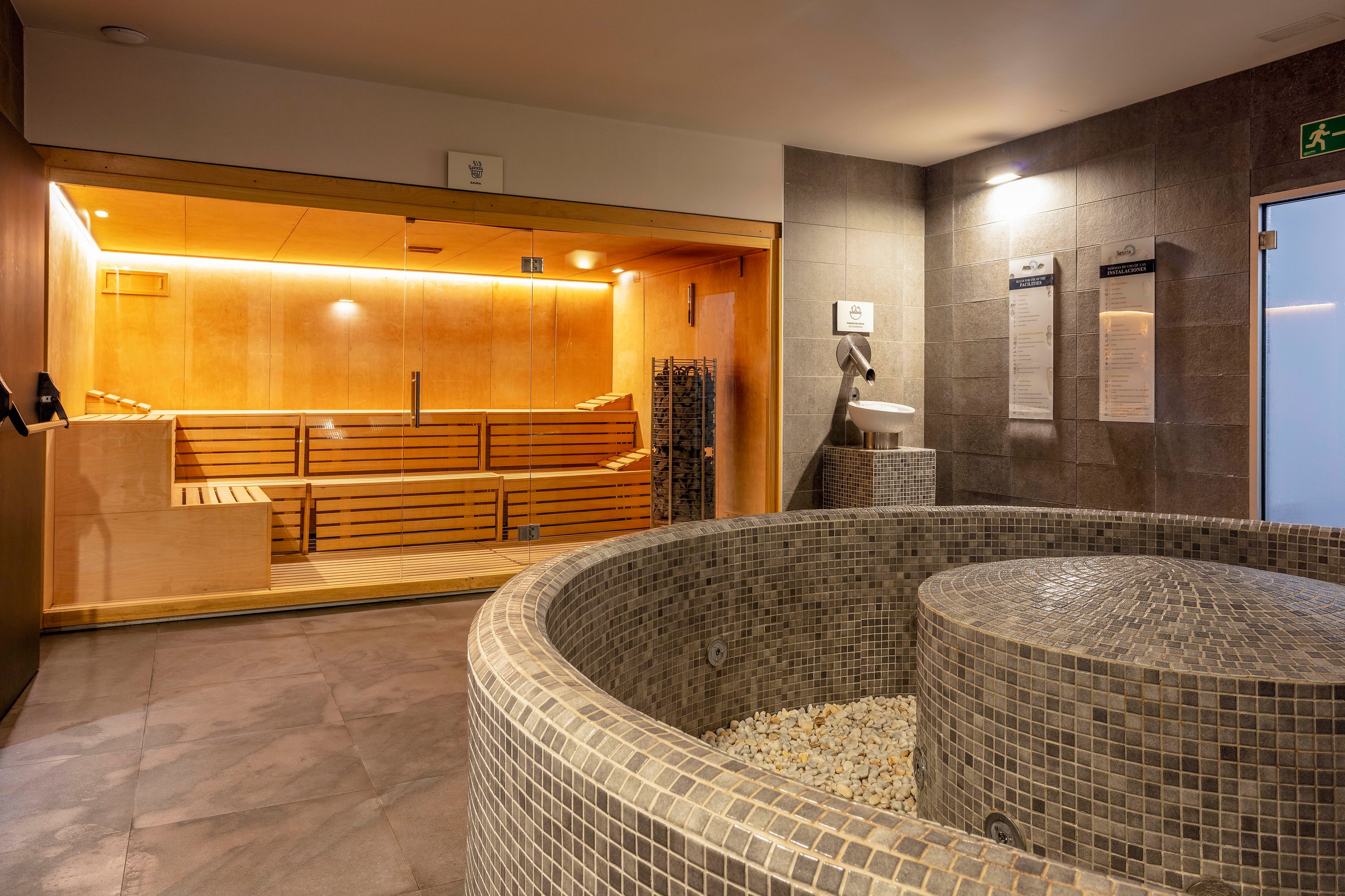 A contemporary spa interior features a wooden sauna room with tiered benches and warm ambient lighting. In the foreground, a round tiled plunge pool or hot tub is filled with stones at the base. The surrounding walls and floor are finished in neutral gray tiles, creating a clean and minimalist atmosphere. Stainless steel fixtures and subtle signage contribute to the modern wellness setting.