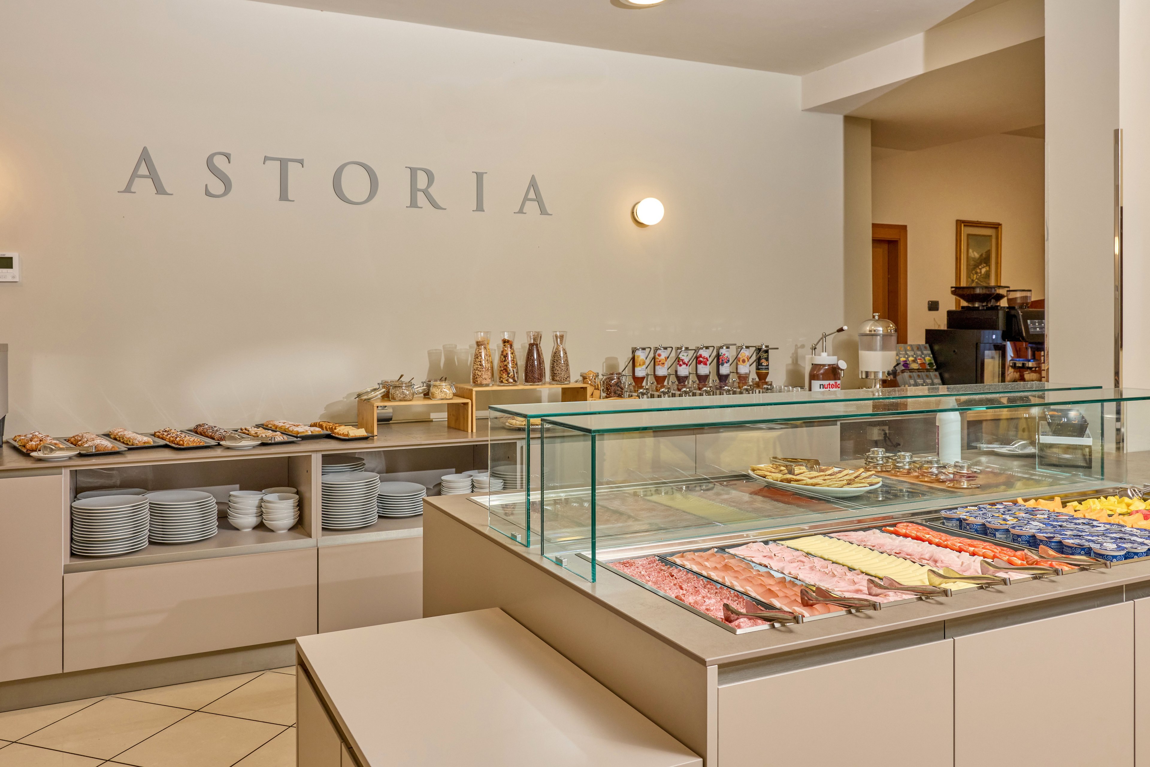 A self-service breakfast buffet is displayed in a hotel dining area with the word ASTORIA on the wall. A glass counter showcases assorted pastries and desserts arranged in neat rows. Behind the counter, shelves hold plates, bowls, and a variety of breakfast items including cereals and baked goods. The space is warmly lit and organized, creating a clean and inviting atmosphere for guests.