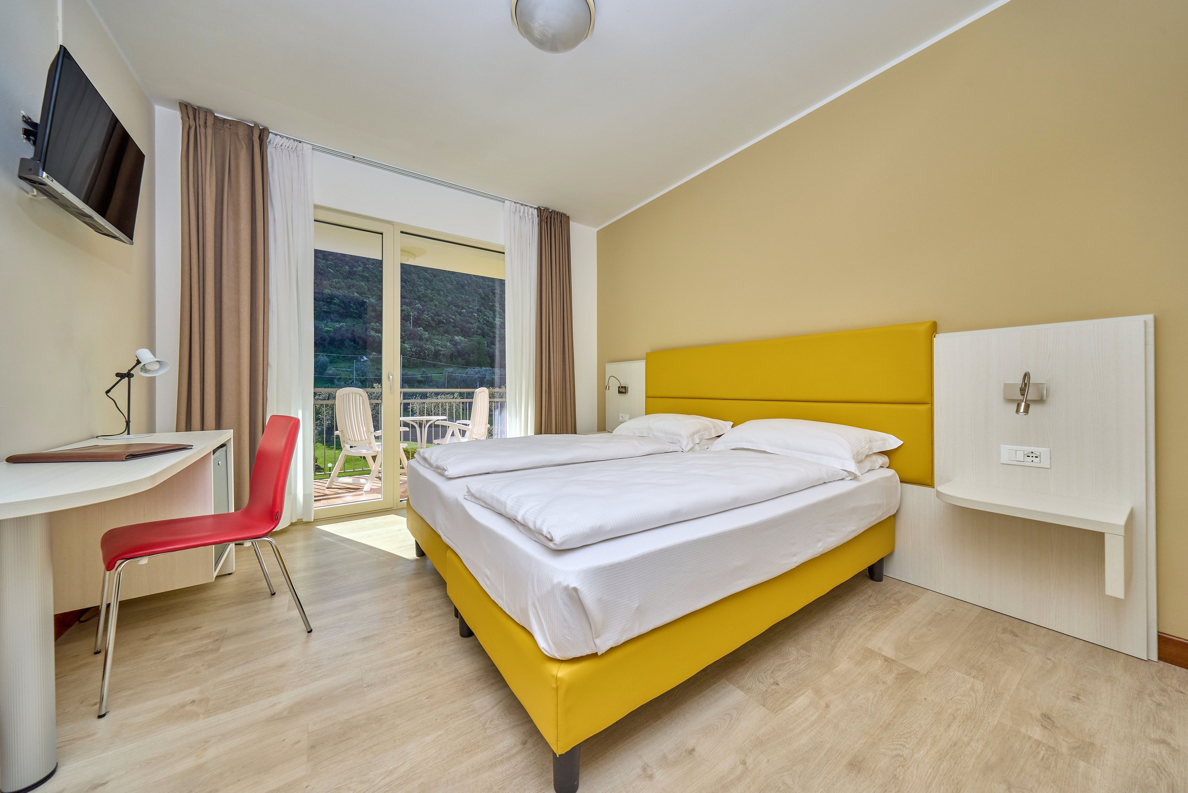 A contemporary hotel room features a double bed with a bold yellow upholstered frame and headboard. The space includes a small white desk with a red chair and a wall-mounted television. Light wood flooring and neutral walls create a clean, minimalist look, complemented by floor-to-ceiling curtains. Glass doors open to a balcony with outdoor seating and a scenic mountain view, allowing natural light to fill the room.