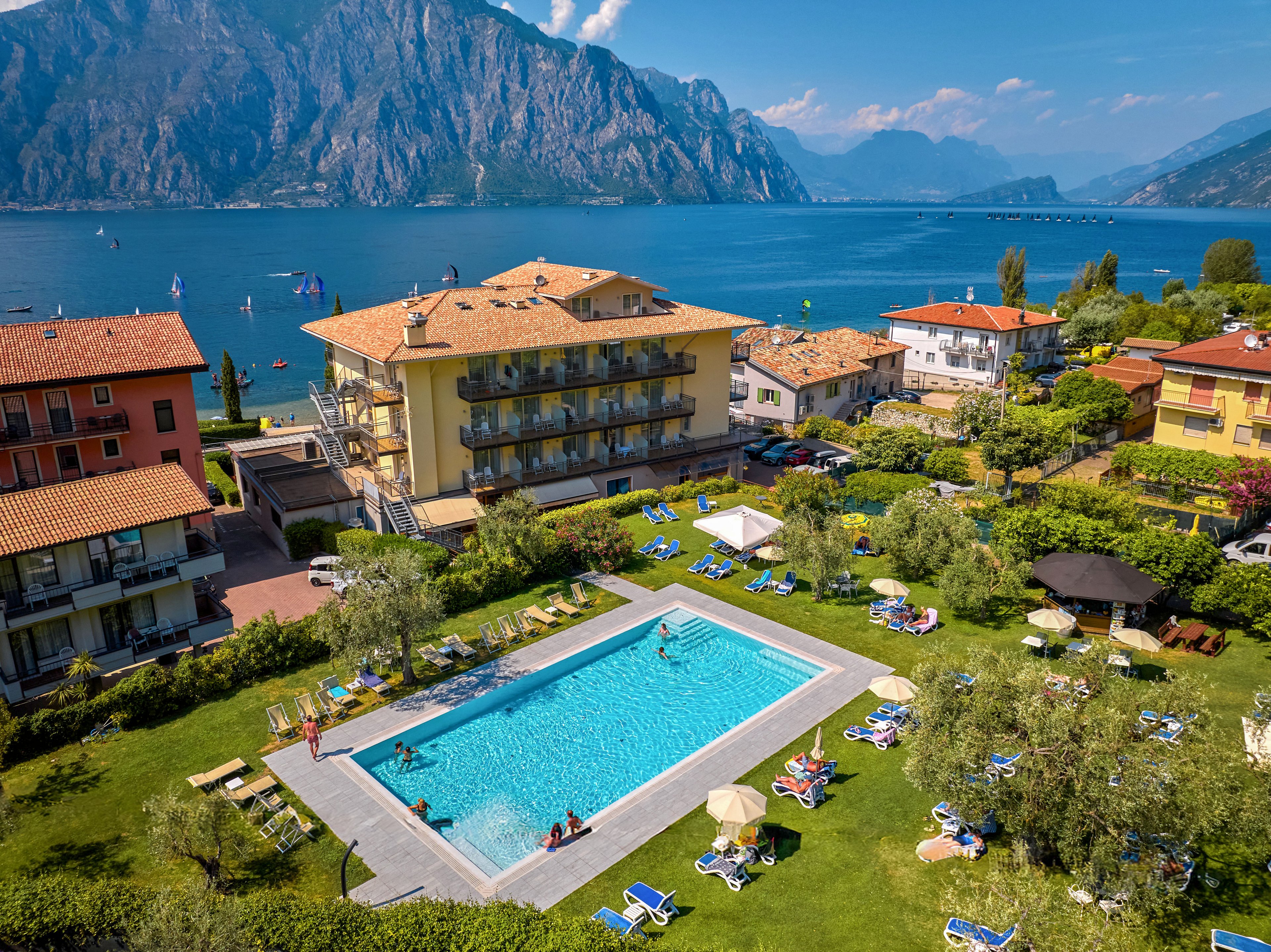 A lakeside hotel complex features a large outdoor swimming pool surrounded by sun loungers and green lawns. Guests relax and swim in the pool while others sunbathe in the garden area. Several multi-story buildings with balconies overlook the water, and sailboats are visible on the lake. Majestic mountains rise in the background under a clear blue sky, creating a scenic vacation atmosphere.