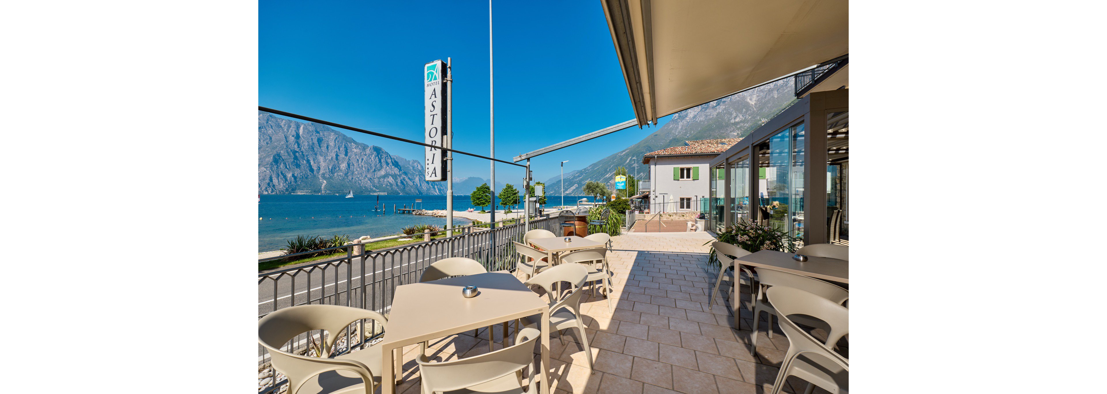 An empty outdoor café terrace overlooks a serene lake with towering mountains in the background. Beige tables and chairs are neatly arranged along a tiled patio beside a metal railing. A vertical sign reading "ASTORIA" rises near the water, and nearby buildings line the waterfront. The scene is bathed in bright sunlight under a clear blue sky, creating a relaxed vacation atmosphere.