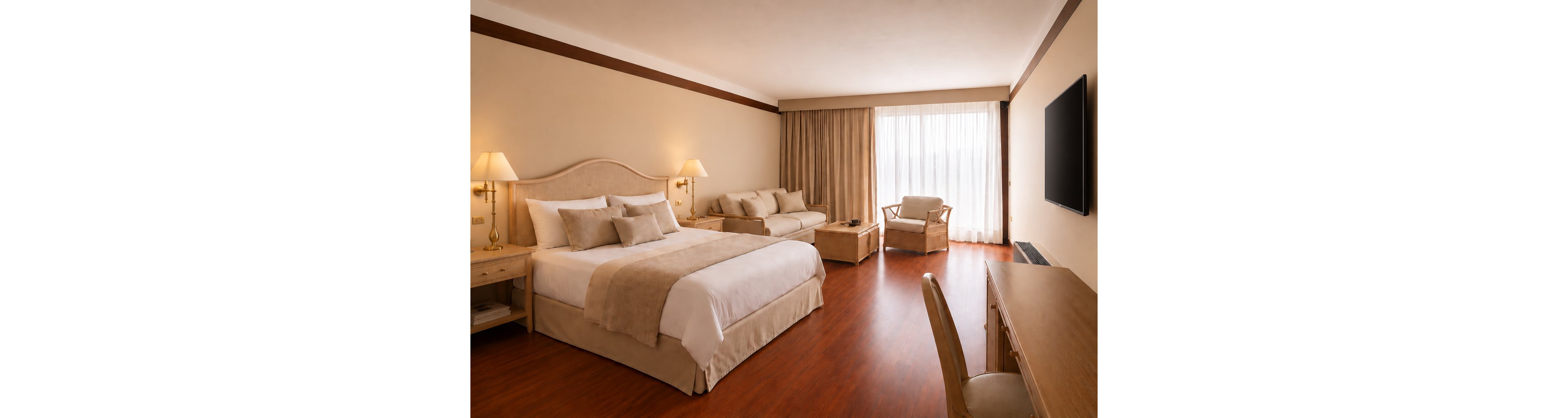 A large, elegant hotel room features a neatly made king-size bed with beige linens and a padded headboard. The space includes a sofa, armchair, and coffee table arranged on warm wooden flooring. Floor-to-ceiling curtains cover a bright window, allowing natural light to fill the room. A flat-screen television is mounted on the wall opposite a wooden desk and chair, creating a comfortable area for work and relaxation.