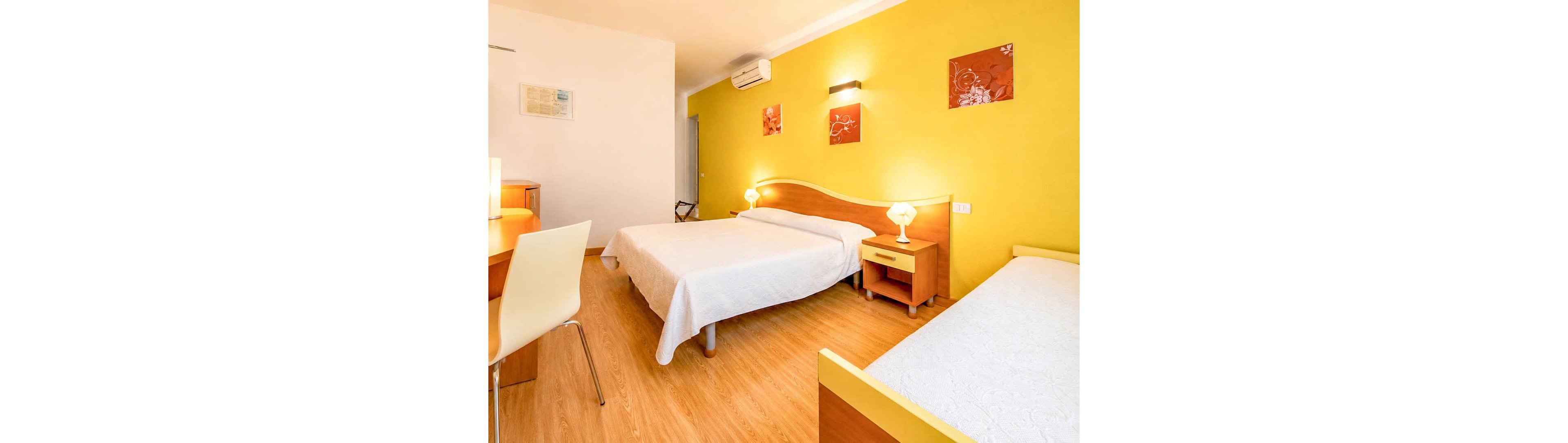 A clean, modern hotel room features a double bed and a single bed with white bedspreads. The space has warm wooden furniture, including bedside tables, a desk, and a chair. A yellow accent wall with floral artwork and wall-mounted lights adds color to the room. Additional details include a table lamp, air conditioner, and framed document on the wall, creating a comfortable and inviting atmosphere.