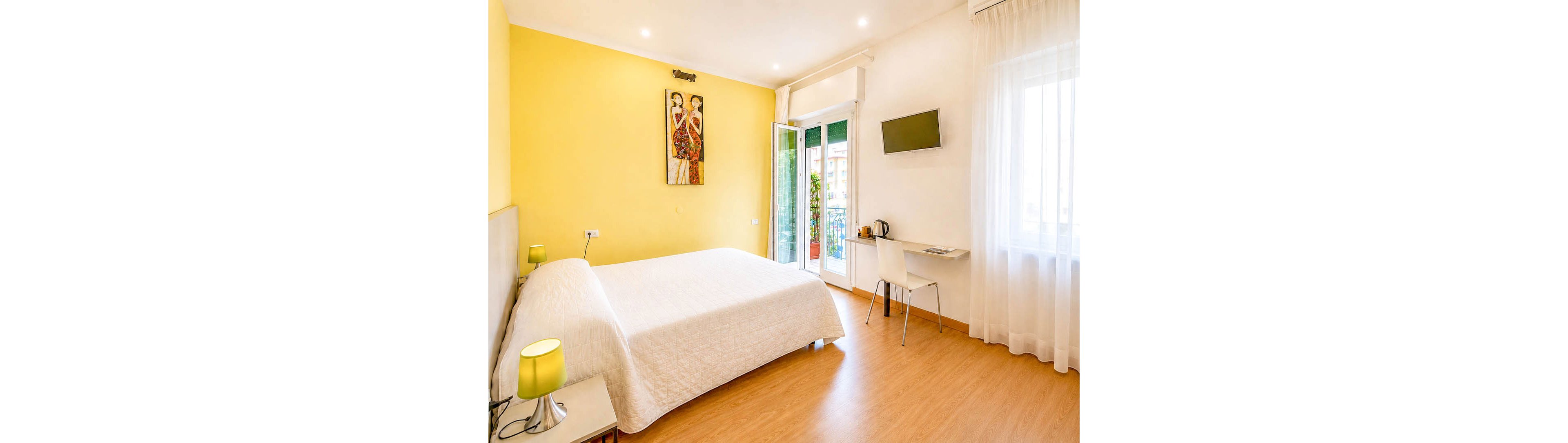 A bright modern bedroom features a large neatly made bed against a yellow accent wall. The room includes a small desk with a chair, electric kettle, and wall-mounted flat-screen TV. Natural light floods in through sheer curtains and glass doors that open to a balcony with plants. Warm wooden flooring and soft lighting create a cozy, inviting atmosphere suitable for guests or travelers.