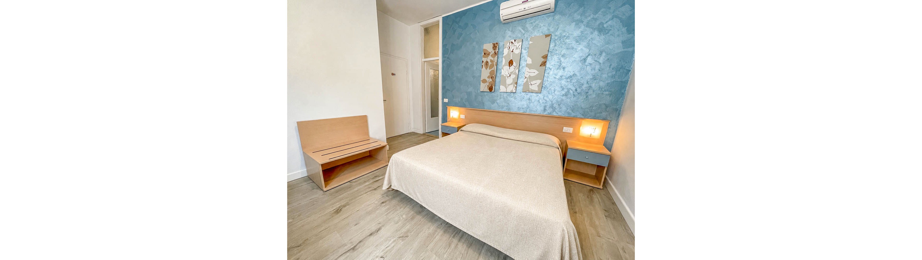 A neatly made double bed with a beige cover stands against a blue accent wall in a minimalist bedroom. Two wooden nightstands with built-in lights flank the bed, creating a warm and inviting atmosphere. Above the headboard, three decorative panels with floral patterns add visual interest. Light wood flooring and simple furniture contribute to the clean, modern design of the room.