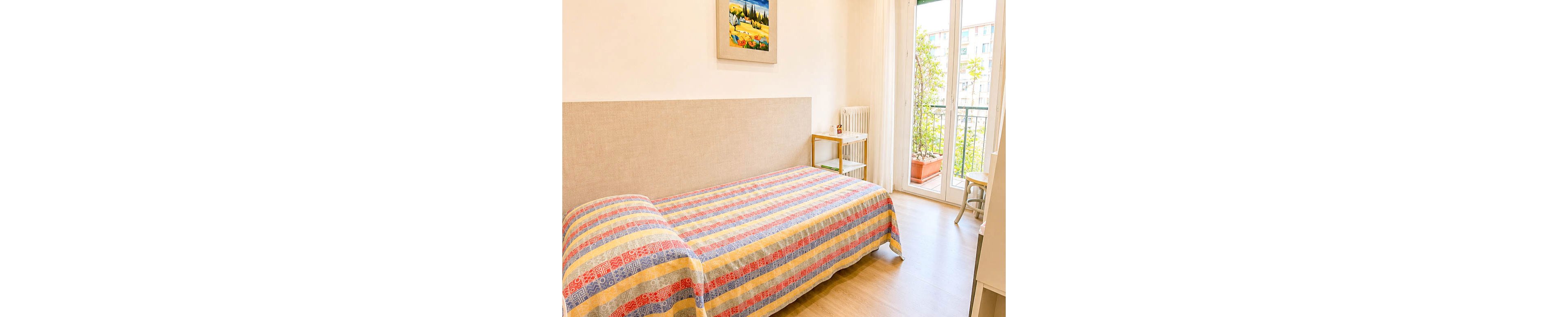 A cozy single bed with a colorful striped bedspread is the main focus of this bright bedroom. The room features light wooden flooring, a simple bedside table, and a radiator against the wall. Large glass doors open onto a balcony with potted plants and a view of nearby buildings. A framed painting adds a decorative touch to the otherwise minimal, sunlit interior.