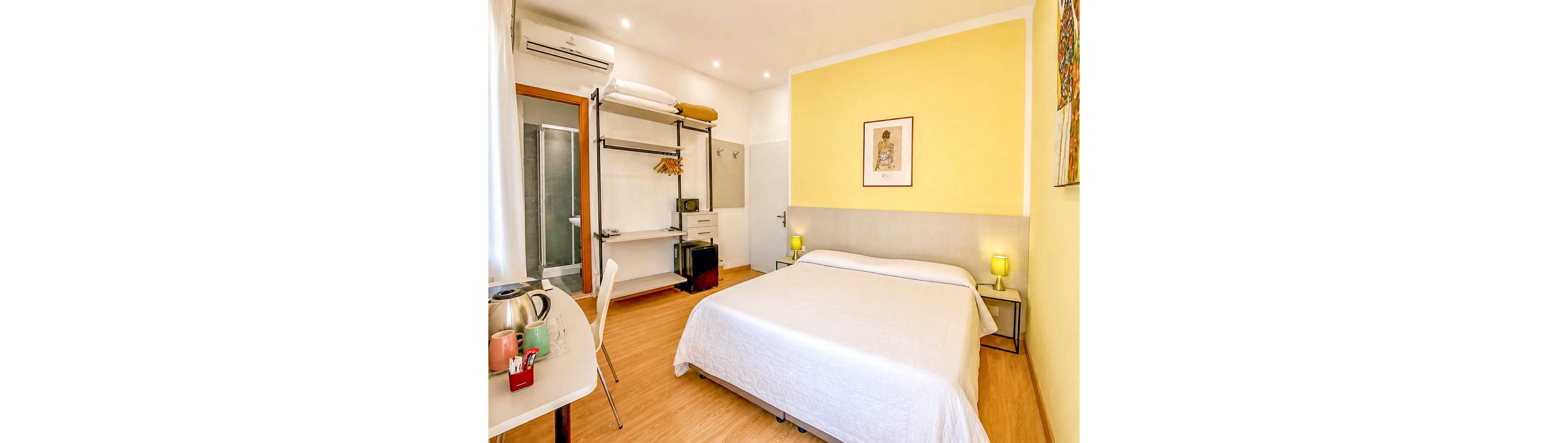 A cozy modern guest bedroom features a neatly made double bed with a white quilt and soft headboard. The room has light yellow accent walls, wooden flooring, and two small bedside tables with matching lamps. An open wardrobe system with shelves, hangers, and a small refrigerator stands near the doorway leading to a glass-enclosed bathroom. A desk with a kettle and cups sits by the window, creating a comfortable and functional stay environment.