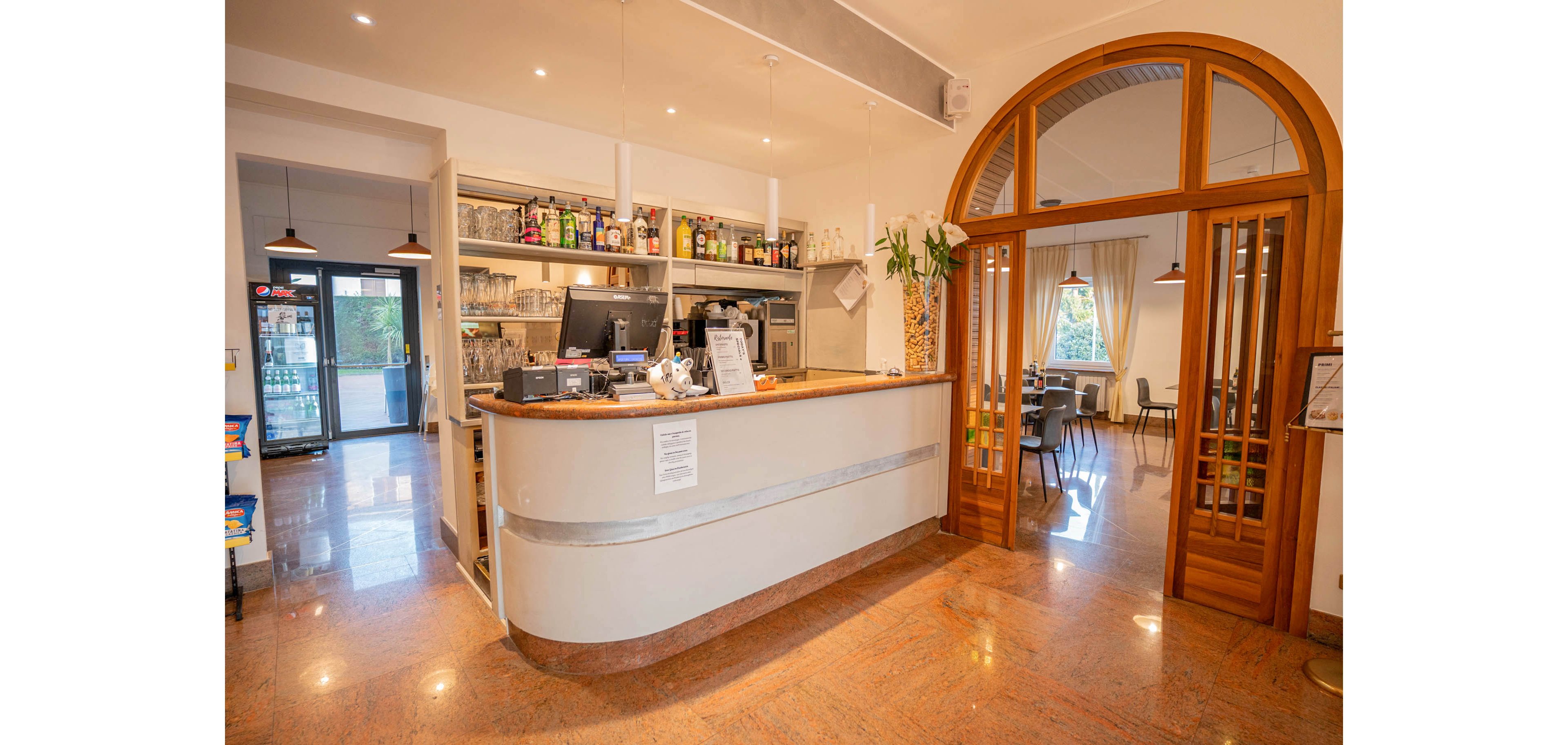 A warm, inviting cafe bar features a curved counter with a computer, coffee machine, and various bottles on shelves behind it. The interior has polished floors, recessed ceiling lights, and neatly organized glassware. A large wooden archway opens into a bright dining room with tables and chairs near tall windows. The overall atmosphere is relaxed and welcoming, suitable for casual drinks or light meals.