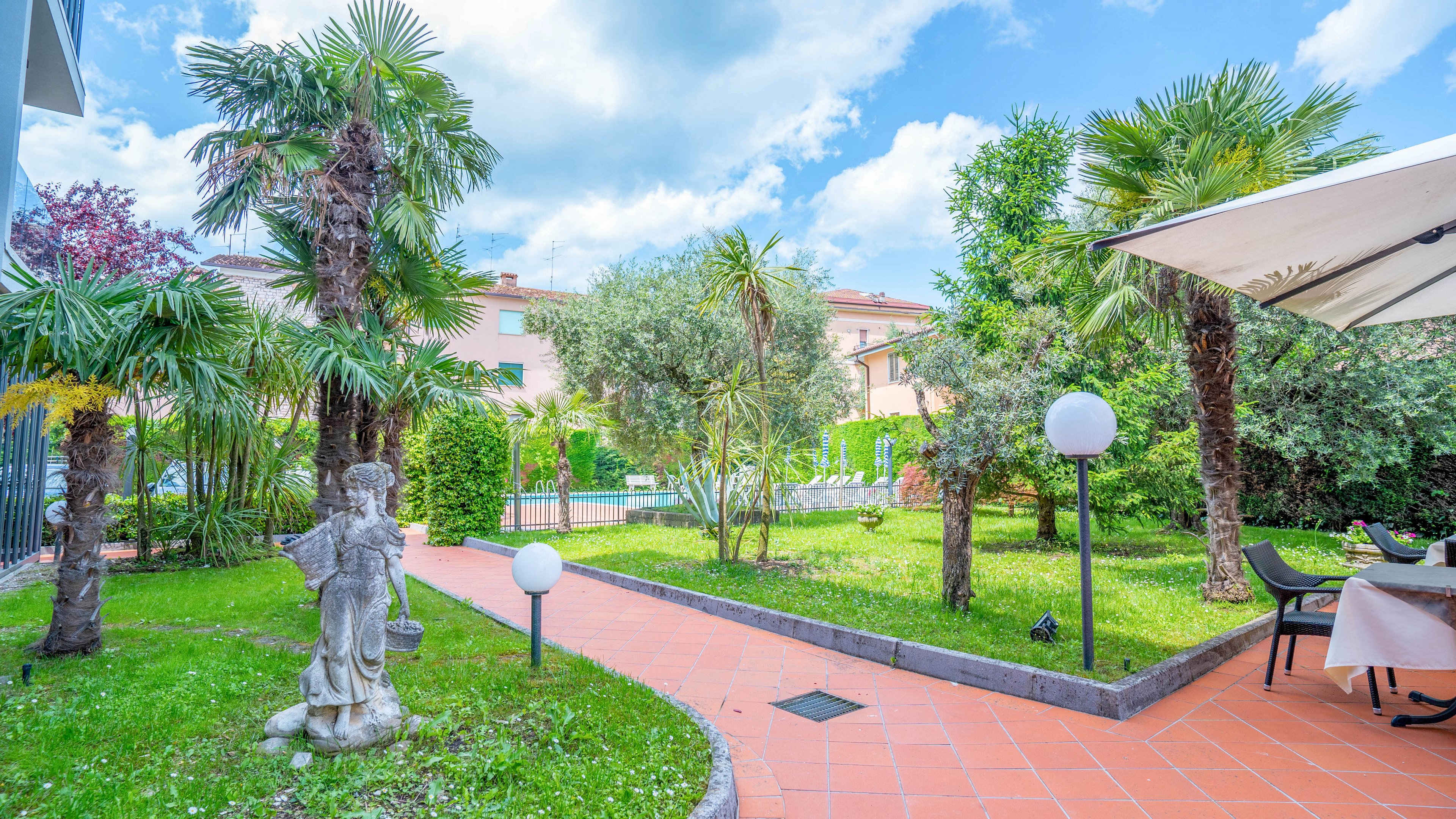 A bright hotel garden features palm trees, manicured grass, and a terracotta-tiled pathway. Decorative statues and round garden lights line the walkway, creating a welcoming outdoor space. In the background, residential-style buildings and a swimming pool area are visible behind hedges. A white patio umbrella shades a small dining table and chairs, suggesting a relaxing spot for guests to sit and enjoy the view.
