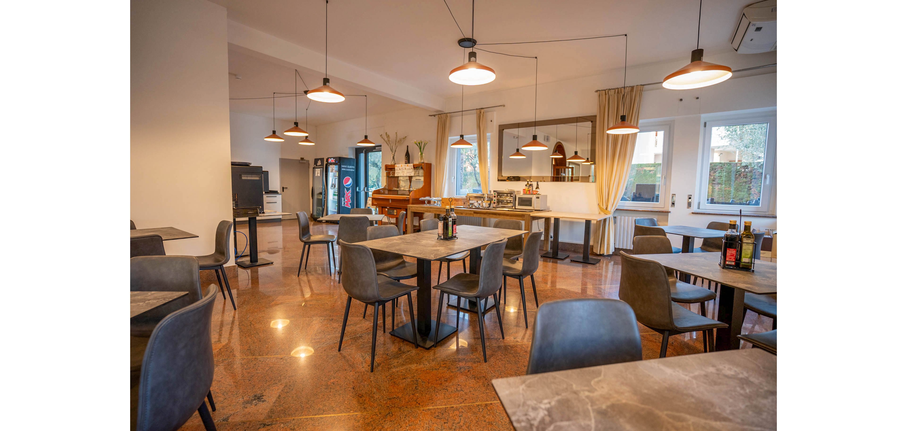 Contemporary restaurant dining room with empty tables and chairs is shown in warm ambient lighting. The space features pendant lights, marble-look tabletops, and polished stone flooring. A buffet or service counter with various items is visible at the back, along with a beverage cooler and large wall mirror. Large windows and glass doors bring in natural light, creating an inviting yet calm atmosphere.
