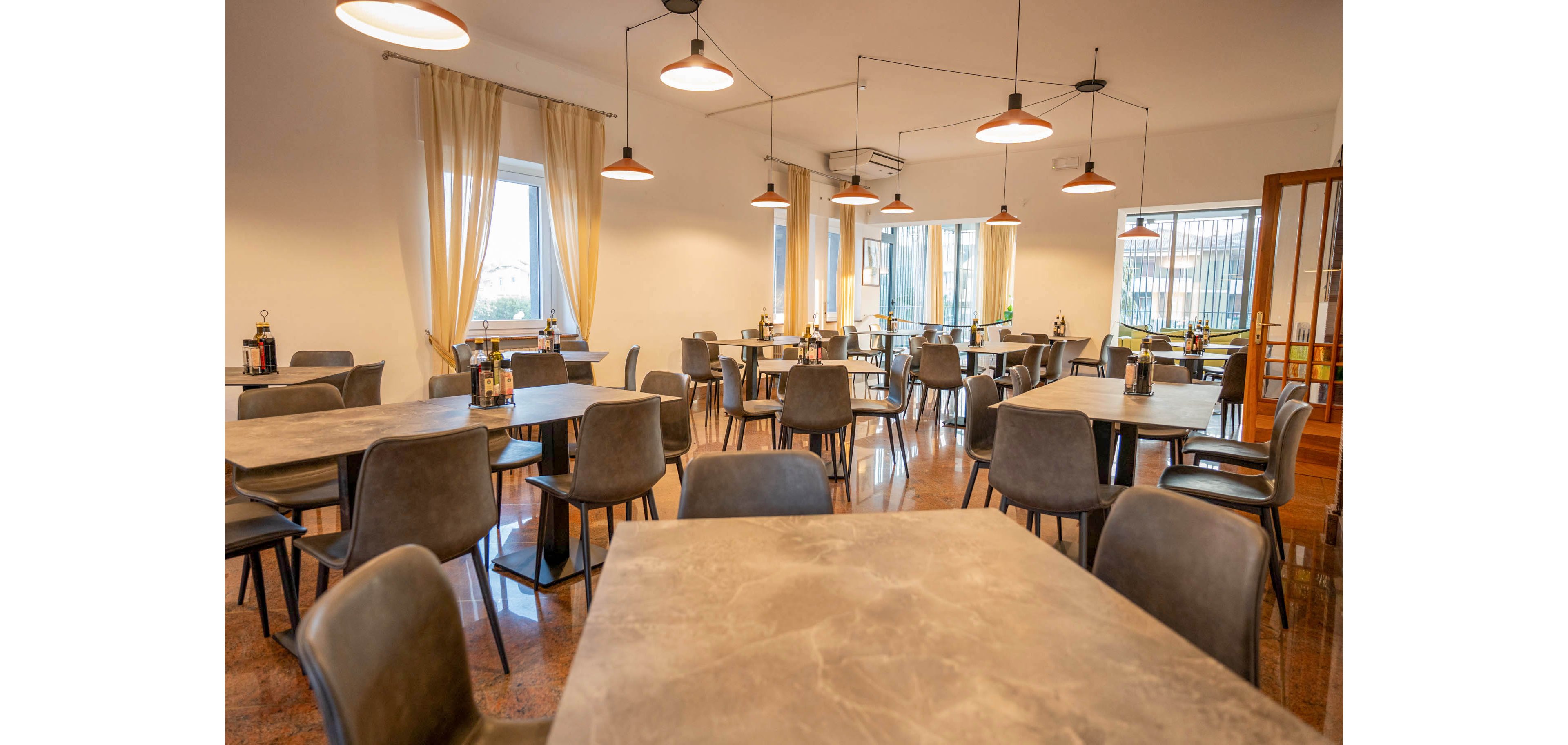 A modern restaurant dining room is shown with multiple empty tables and chairs neatly arranged. Warm pendant lights hang from the ceiling, casting a soft glow over the polished floor. Large windows with light curtains allow natural daylight to brighten the space. Bottles and condiments are placed on the tables, suggesting the room is prepared for upcoming guests.