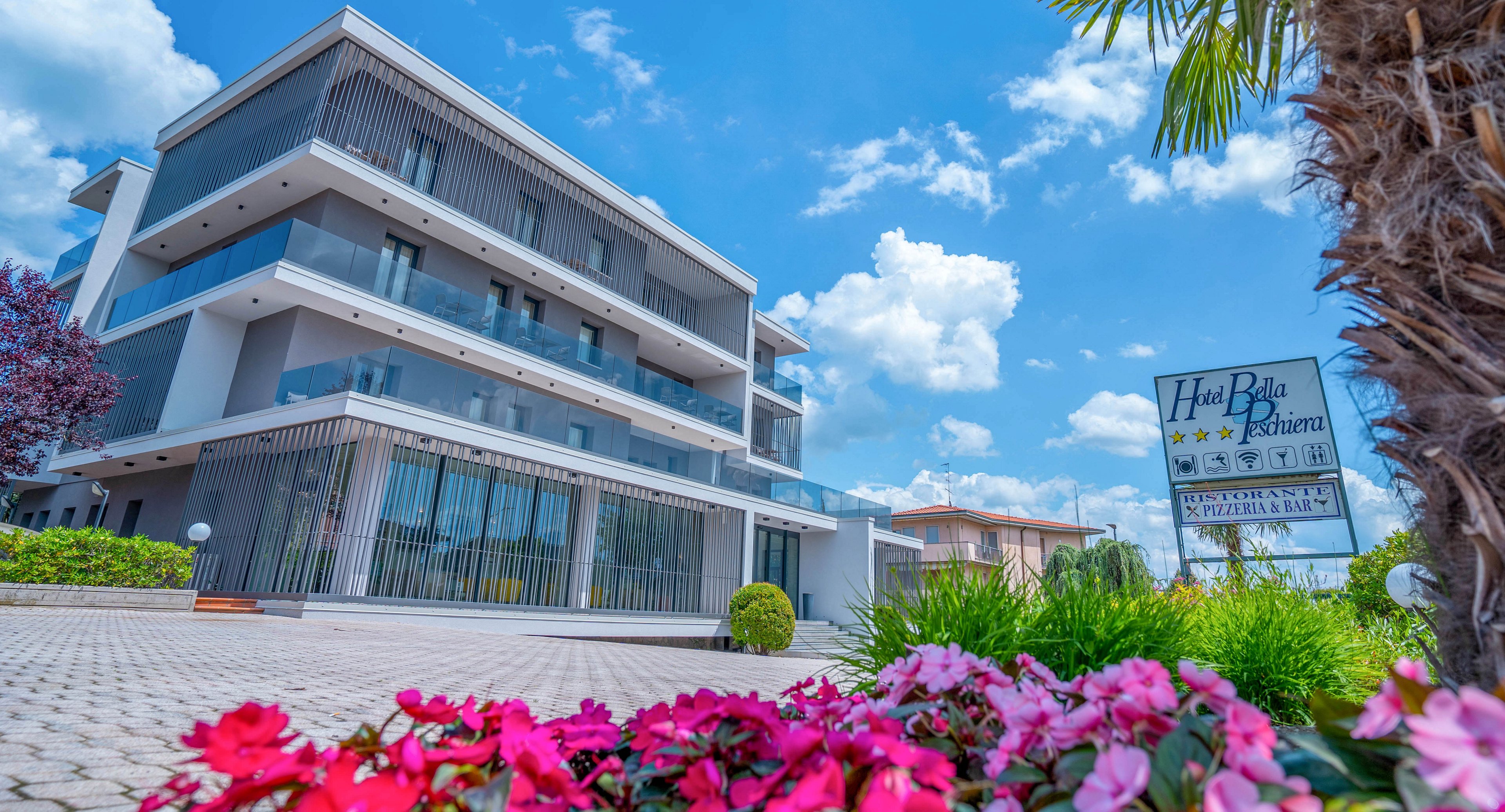 A contemporary multi-story hotel building is shown from a low angle on a bright, sunny day. The foreground features vivid pink flowers and green shrubs leading toward the entrance. A signboard near the street reads "Hotel Bella Riviera" with three stars clearly visible. The sky is blue with scattered white clouds, creating a welcoming vacation atmosphere.