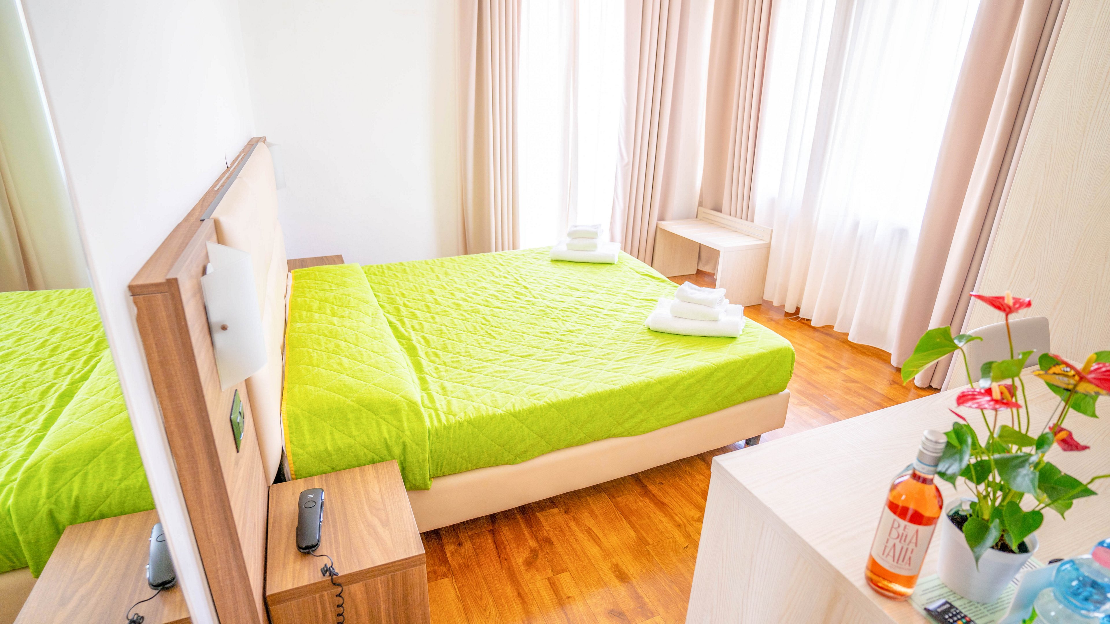 A tidy bedroom features a double bed with a bright green quilt and neatly folded white towels on top. The room has light wood furniture, including bedside tables and a desk, set on a warm wooden floor. Large windows with beige curtains allow natural light to fill the space. On the desk, there are bottled drinks and a potted plant adding color and a welcoming touch.
