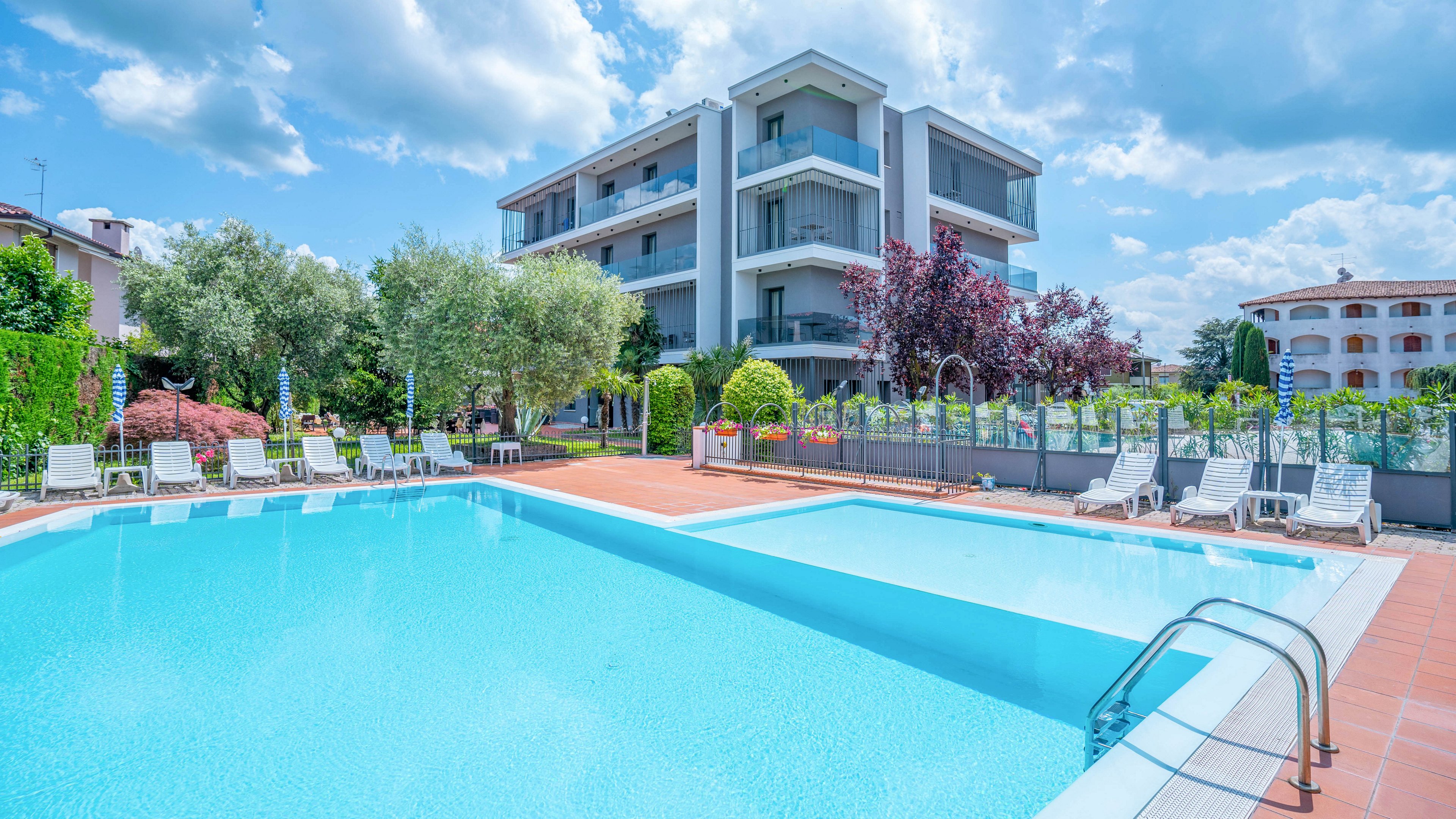 A large outdoor swimming pool is situated in front of a modern multi-story apartment building. White lounge chairs and umbrellas line the poolside terrace, suggesting a relaxing leisure area. The surrounding landscaping includes trees, shrubs, and a neatly maintained fence. A bright blue sky with scattered clouds enhances the summery, vacation-like atmosphere.