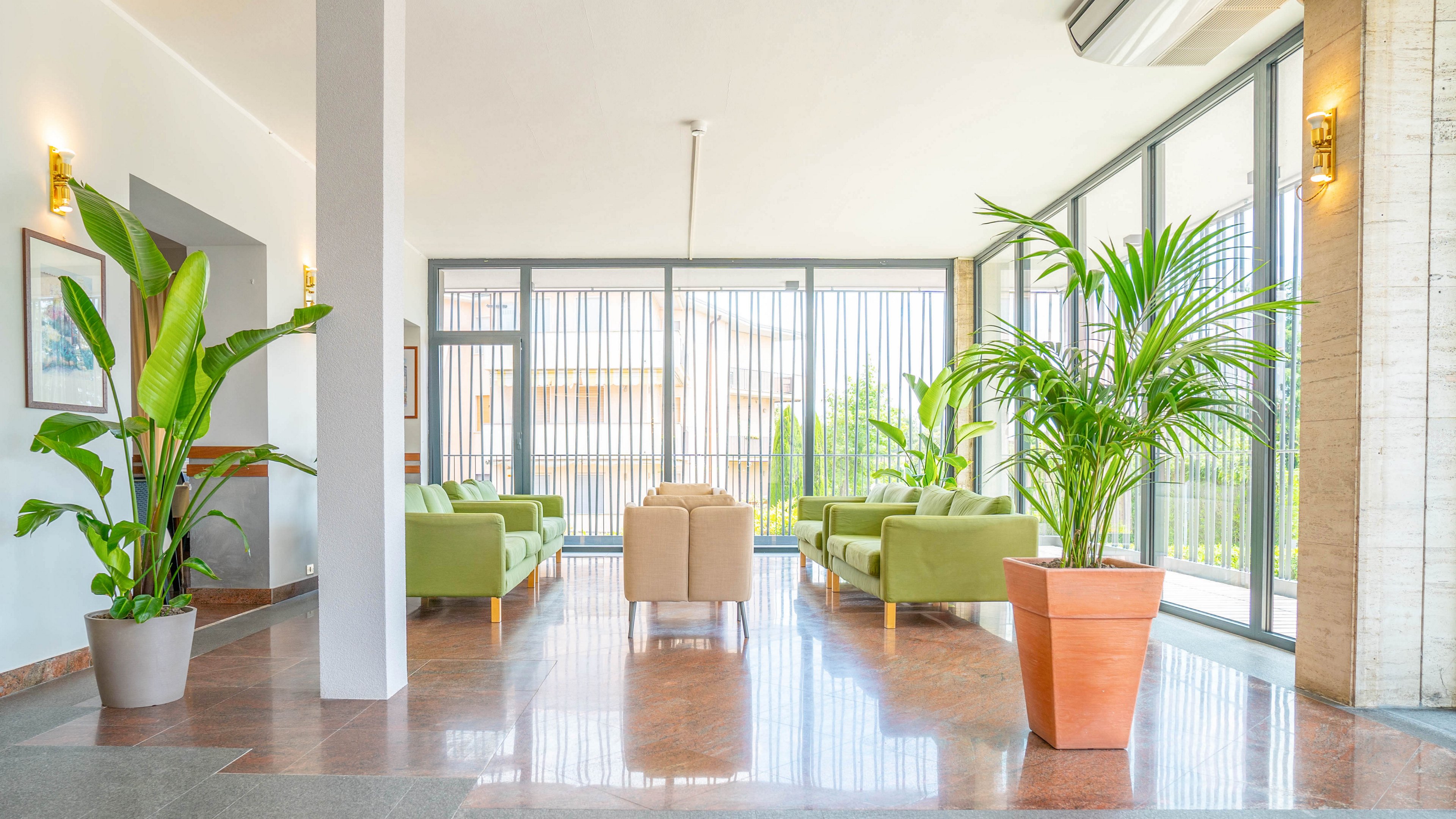 A spacious, modern lobby features several green sofas arranged around a central beige armchair set. Large floor-to-ceiling windows line one side of the room, allowing abundant natural light to flood the space. Potted tropical plants add greenery and contrast against the polished stone floor and neutral walls. Wall sconces and framed artwork contribute to the contemporary, welcoming atmosphere.