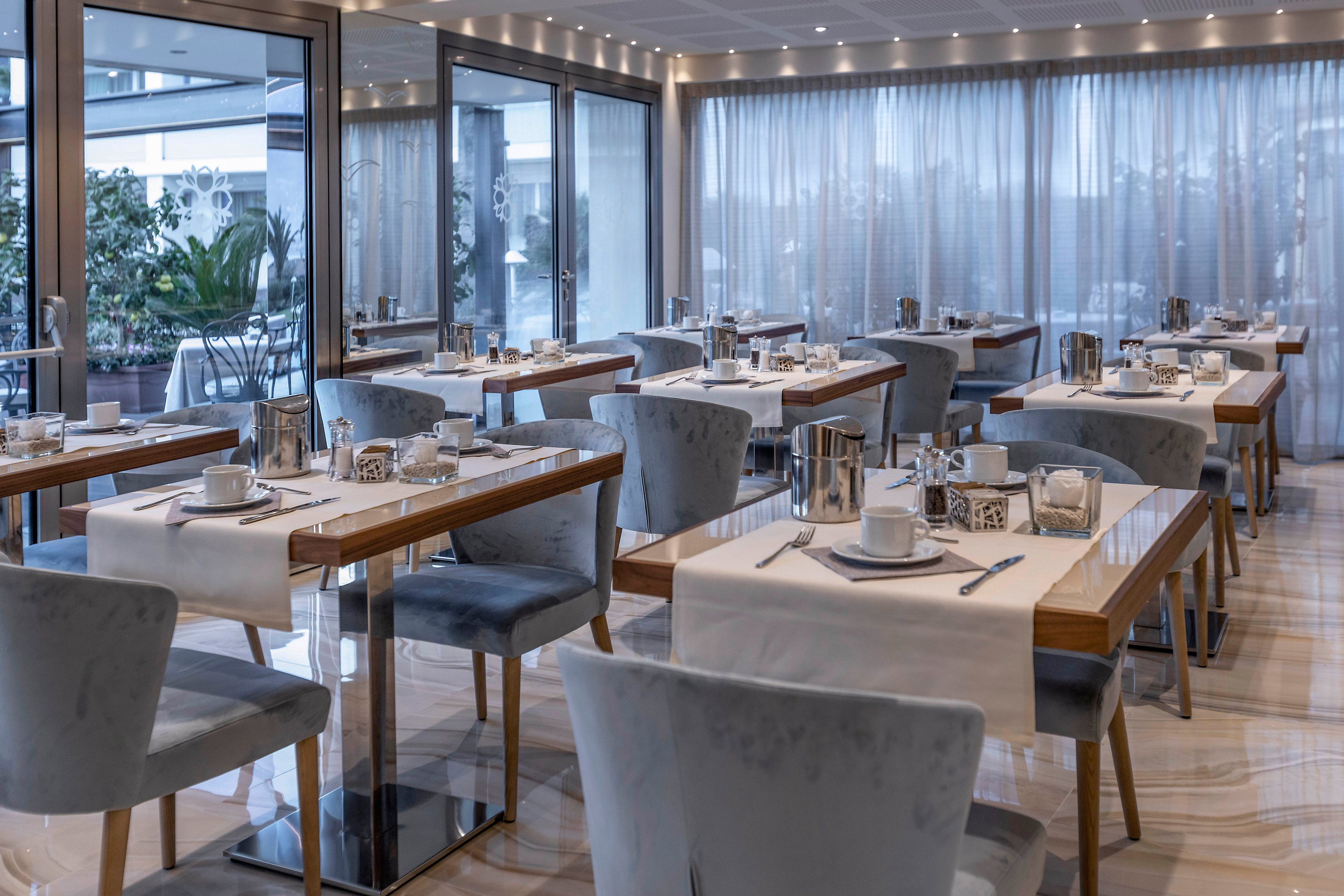 A contemporary restaurant dining area is neatly arranged with wooden tables and soft gray upholstered chairs. Each table is set with white table runners, cups, saucers, cutlery, and small decorative items, ready for breakfast service. Large windows and glass doors let in natural light and reveal greenery on the terrace outside. Sheer curtains, recessed ceiling lights, and polished floors create a calm, elegant atmosphere suitable for hotel guests or business travelers.