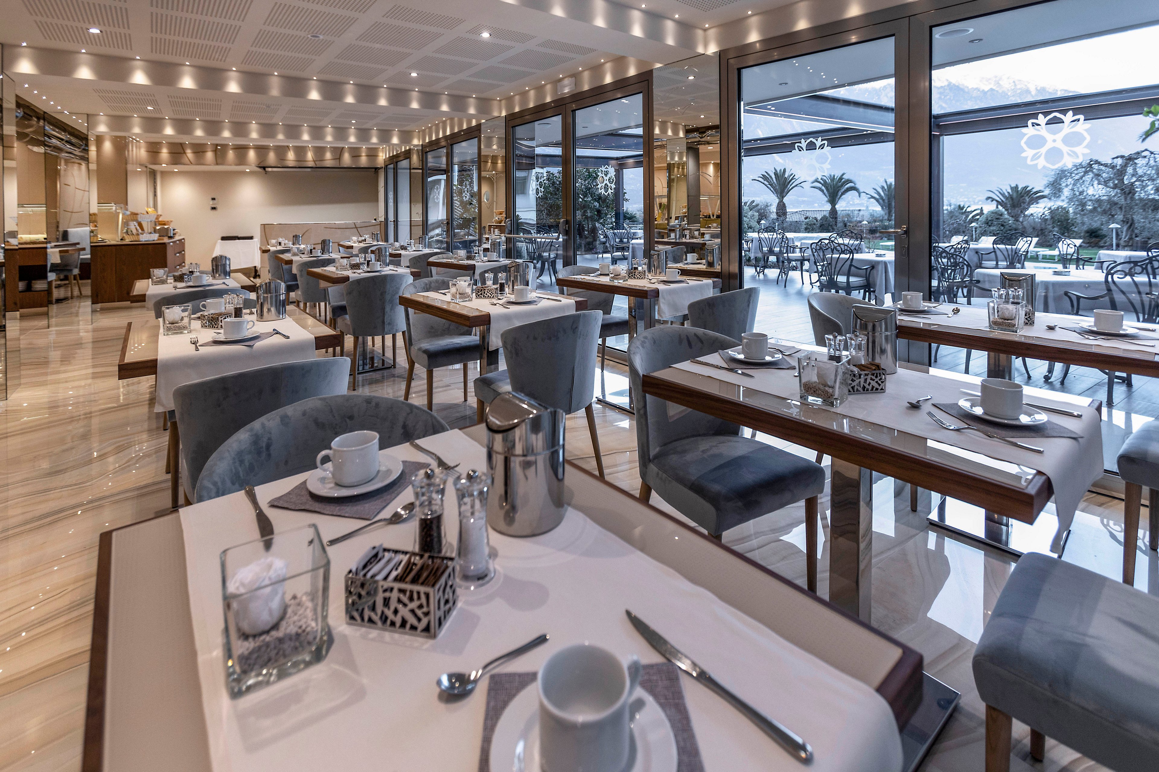 A modern hotel restaurant is neatly arranged with tables set for breakfast. White tableware, metal coffee pots, and glassware are placed on crisp placemats atop wooden tables. Large windows open to an outdoor terrace with additional seating and palm trees in the background. Soft ceiling lights and reflective surfaces create a calm, upscale dining atmosphere.