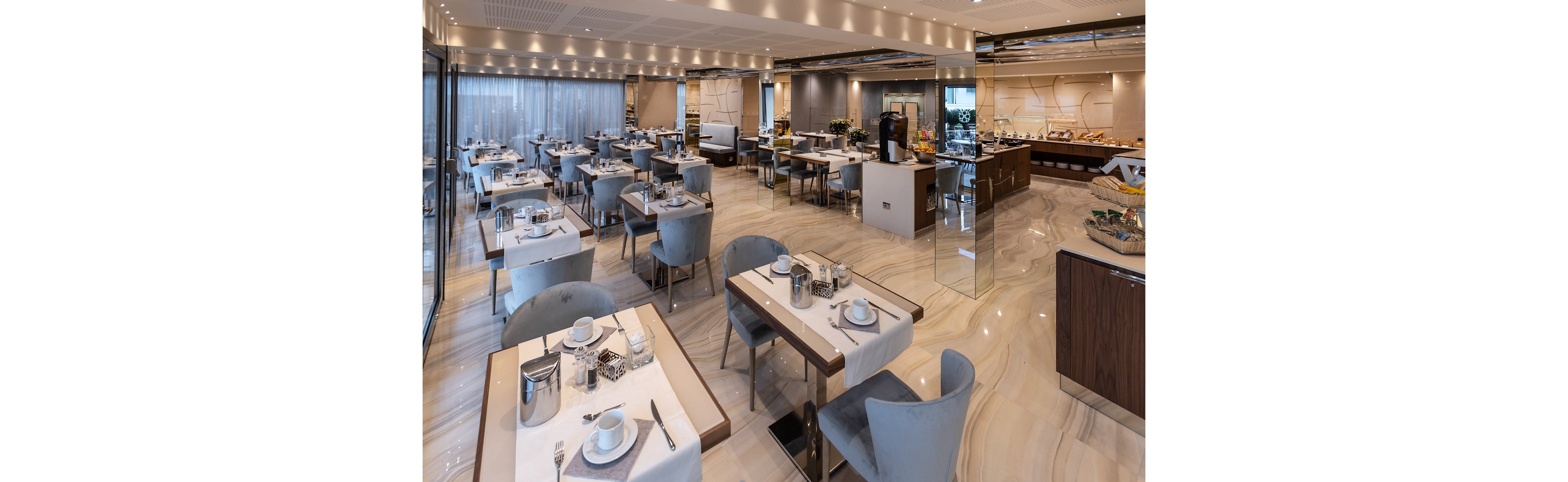 Spacious contemporary restaurant dining room set for breakfast with neatly arranged tables and chairs. The interior features polished marble floors, soft gray seating, and large windows with sheer curtains along one side. Place settings include white dishes, cups, and metal coffee pots on light table runners. In the background, a buffet counter displays various food items under warm ceiling lighting, creating a clean and inviting atmosphere.