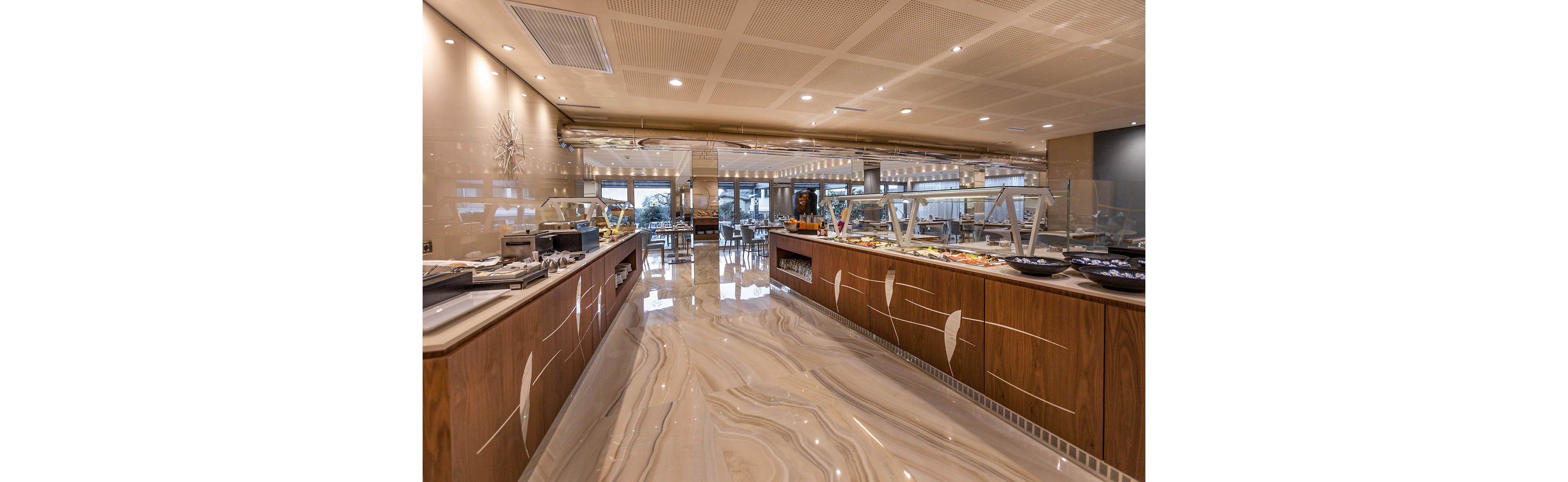 A spacious, modern buffet restaurant features long wooden counters with glass-covered food stations. Various dishes and salads are neatly arranged along the self-service line. The polished marble-style floor and warm ceiling lighting create a clean, upscale atmosphere. In the background, dining tables and chairs are set for guests in a bright, open seating area.