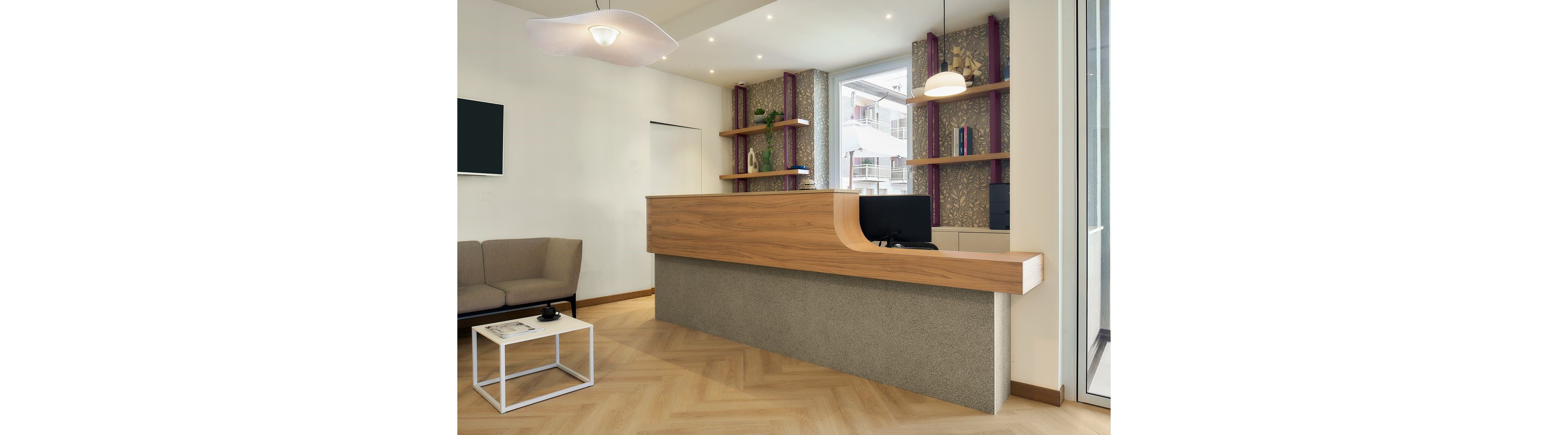 A contemporary reception desk with a wood and stone finish stands in a bright office lobby. Shelving with decorative items and patterned panels lines the back wall, while large windows reveal an urban street outside. A small seating area with a sofa and minimalist coffee table is positioned to the side. The space is illuminated by recessed ceiling lights and pendant lamps, creating a clean and welcoming atmosphere.