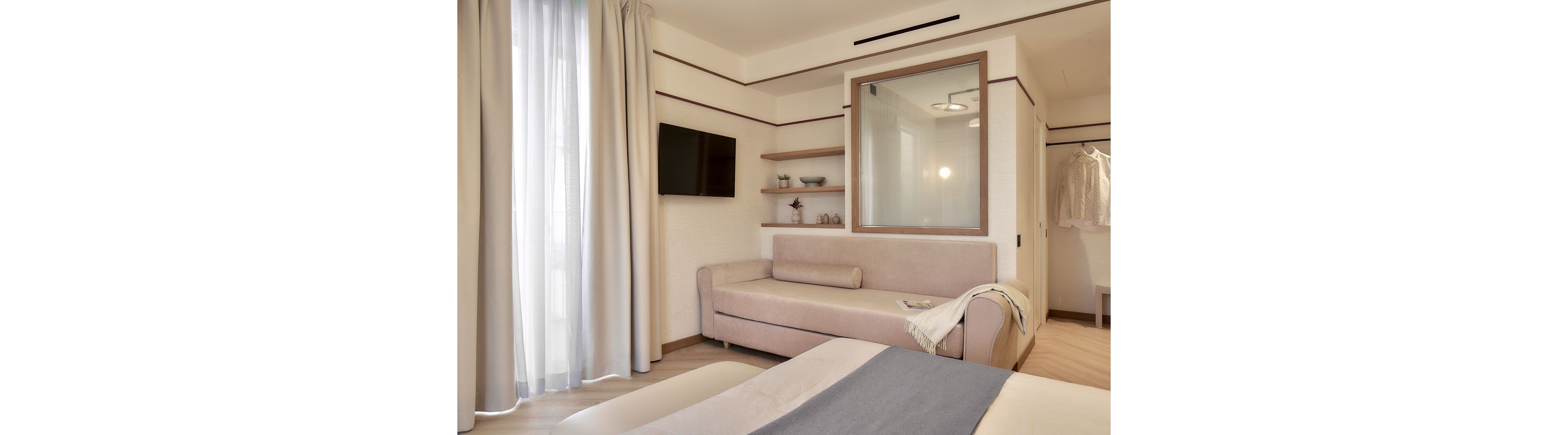 A contemporary hotel room features a beige sofa with cushions and a soft throw draped over the arm. A flat-screen TV is mounted on the wall opposite sheer curtains that let in natural light. Built-in wooden shelves display minimal decorative objects beside a large interior window. Neutral tones and clean lines create a calm, inviting atmosphere suitable for relaxation or business travel.