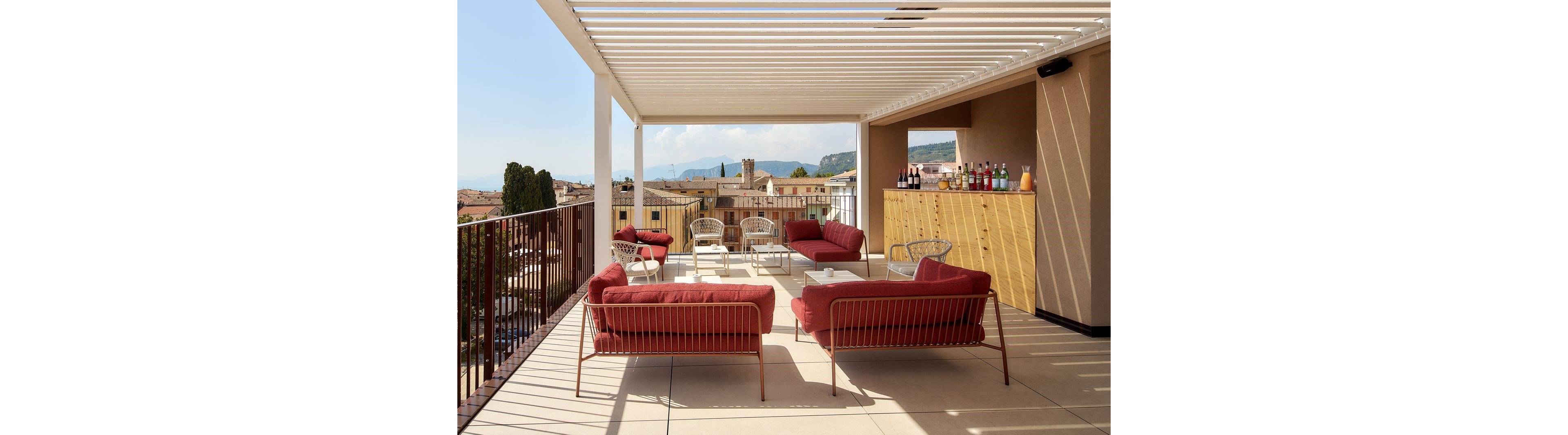 An open rooftop terrace features modern red cushioned sofas and metal chairs arranged for social seating. A wooden bar with assorted bottles stands along one side under a white pergola structure. The terrace overlooks a picturesque town with historic buildings, trees, and distant hills under a bright sky. The scene suggests a relaxed outdoor gathering space with contemporary design and warm, inviting colors.