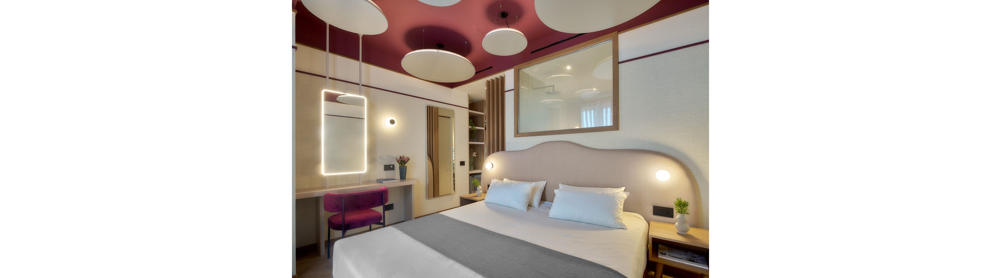 Contemporary hotel room featuring a neatly made double bed with white linens and a gray throw. The space includes a work desk with a purple chair, illuminated by a vertical rectangular mirror light. A wardrobe and open shelving unit with decor items are visible near the bed. The ceiling showcases bold circular panels in white and burgundy, reflected in a large interior window above the headboard. Soft ambient lighting and minimal decor create a cozy, modern atmosphere.