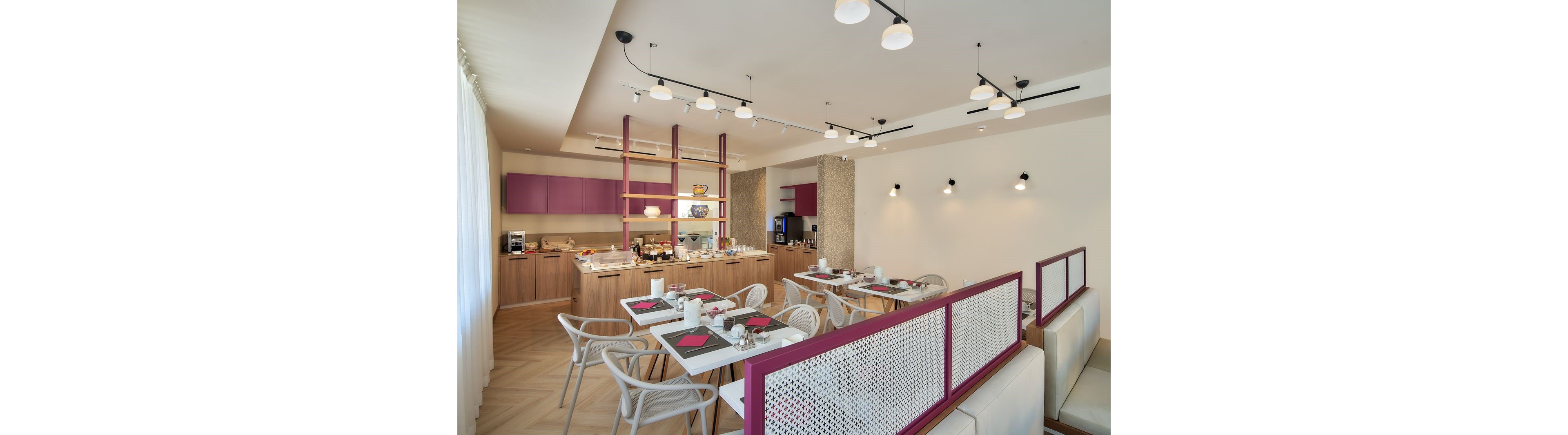 Contemporary dining room with neatly arranged tables in a hotel or café setting. White chairs and tables are accented with purple details and red placemats, creating a clean, modern look. A buffet counter with food, drinks, and coffee machines lines the back wall beneath purple cabinets. Warm lighting and light wood flooring contribute to a bright, welcoming atmosphere.