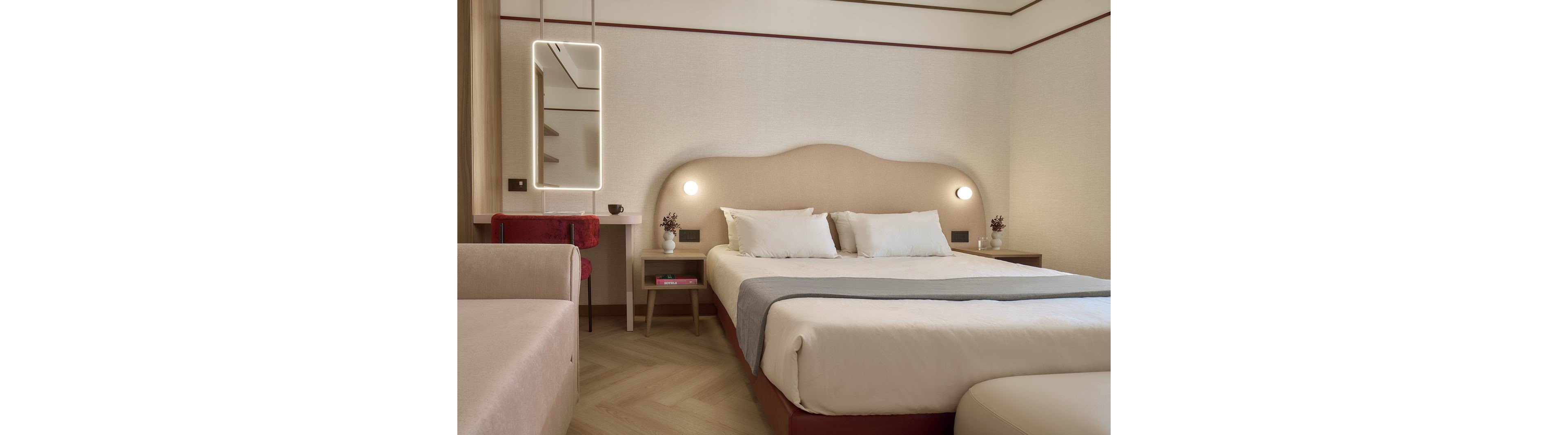 A contemporary hotel bedroom features a large double bed with crisp white linens and a soft beige headboard. The room includes matching bedside tables with small lamps and decorative plants. A lighted wall mirror and a red chair add subtle color accents to the otherwise neutral palette. Soft, indirect lighting and clean lines create a calm, minimalist atmosphere suitable for relaxation.