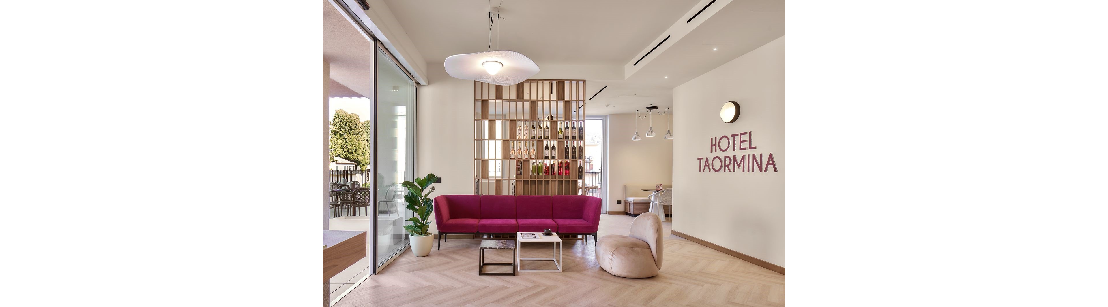 Contemporary hotel lobby interior featuring a bright pink sofa and a beige lounge chair. A wooden partition with shelves displaying bottles separates the seating area from the rest of the space. Large glass doors open to an outdoor terrace, allowing natural light to fill the room. The wall text reads "HOTEL TAORMINA," and the decor includes minimalist lighting and a potted plant.