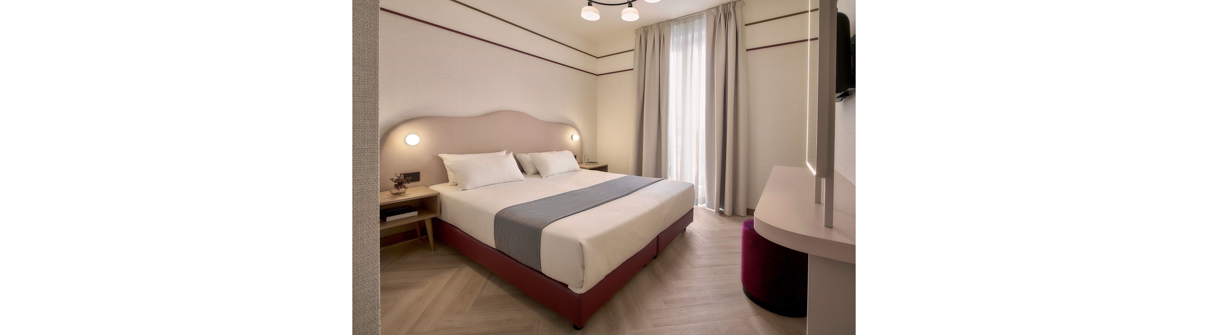 A contemporary hotel room features a neatly made double bed with white linens and a gray runner. Soft wall-mounted lights flank the upholstered headboard, creating a warm and inviting atmosphere. Light wood flooring and neutral-colored walls complement the floor-to-ceiling curtained window. A minimalist desk and a round upholstered stool complete the clean, modern interior design.