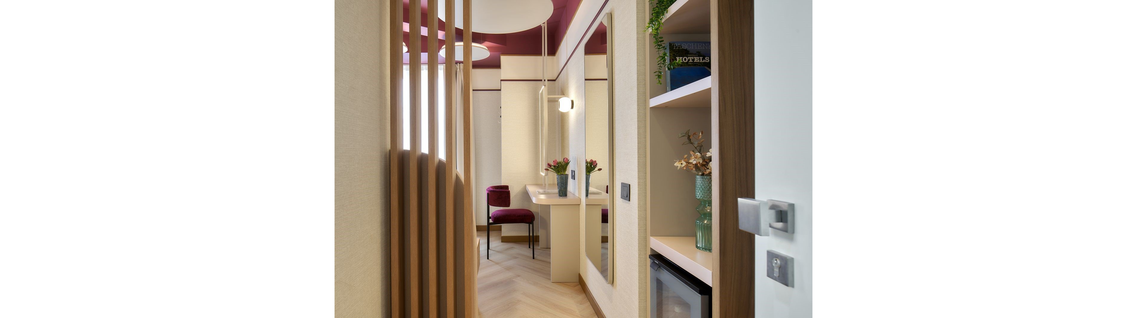 A narrow, contemporary hotel room corridor leads to a vanity desk with a mirror and a cushioned chair. Soft beige walls and wooden slats contrast with a deep burgundy ceiling and accents. A small refrigerator is built into a shelving unit that also holds decorative plants and a book titled "HOTELS". Warm lighting and fresh flowers on the desk create a cozy, inviting atmosphere.