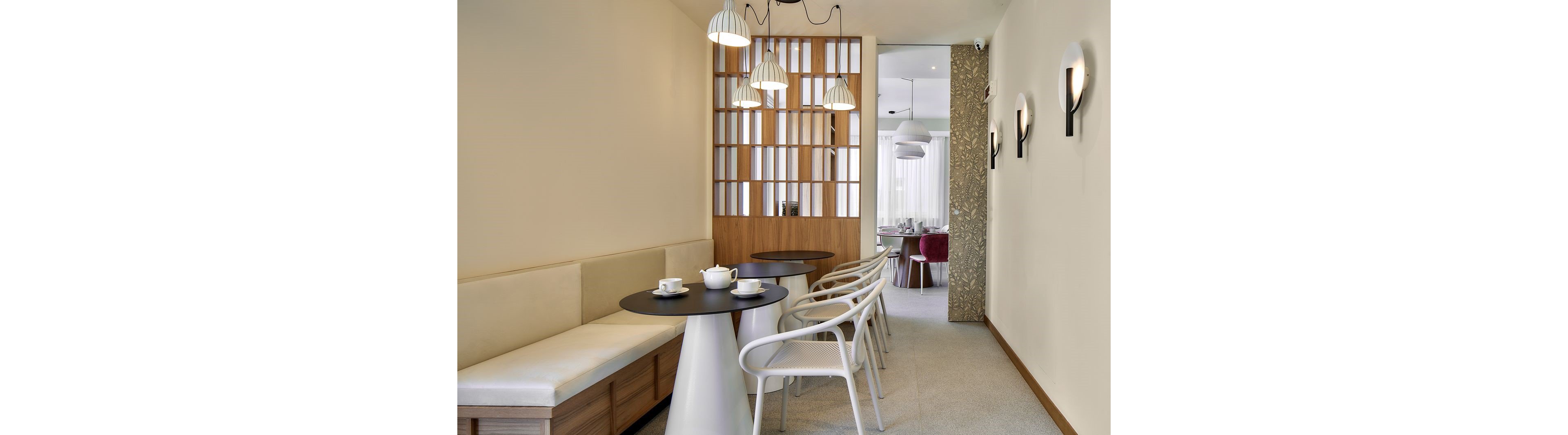 Contemporary café interior featuring round black tables, white chairs, and a built-in bench along a light-colored wall. Pendant lights hang above the seating area, complemented by wall sconces that add soft ambient lighting. A wooden slatted partition separates the foreground from a secondary dining space with red upholstered chairs. The overall design is clean and minimalist, with neutral tones and subtle textures creating a calm, inviting atmosphere.