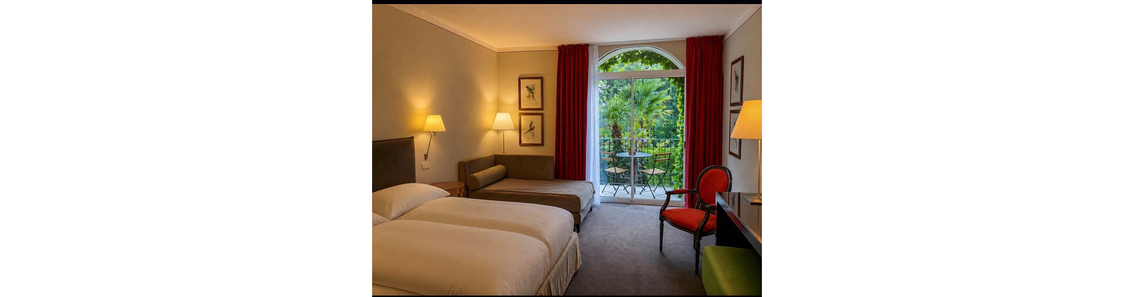 A contemporary hotel room features two beds, a chaise lounge, and a desk with a red chair. The space is warmly lit with wall lamps and decorated with framed bird artwork. Large glass doors open to a balcony overlooking lush greenery and outdoor seating, creating a tranquil atmosphere.