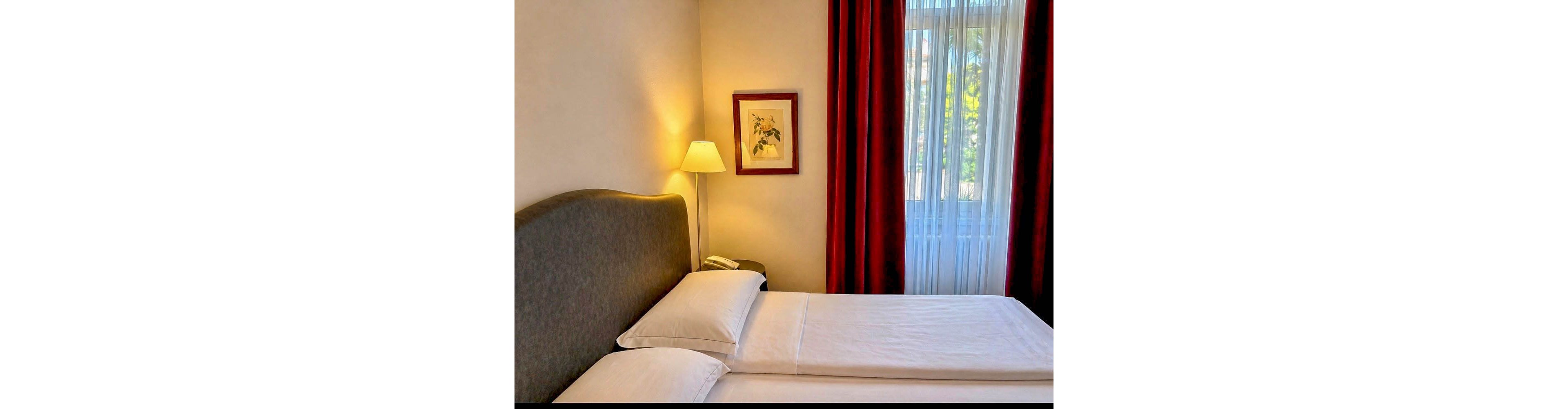 A neatly arranged hotel room features two twin beds with crisp white linens and a padded headboard. The space is illuminated by a standing lamp beside a framed floral artwork, and natural light streams in through a large window with sheer curtains and deep red drapes. The overall mood is calm and inviting, with a neutral palette and minimal decor.