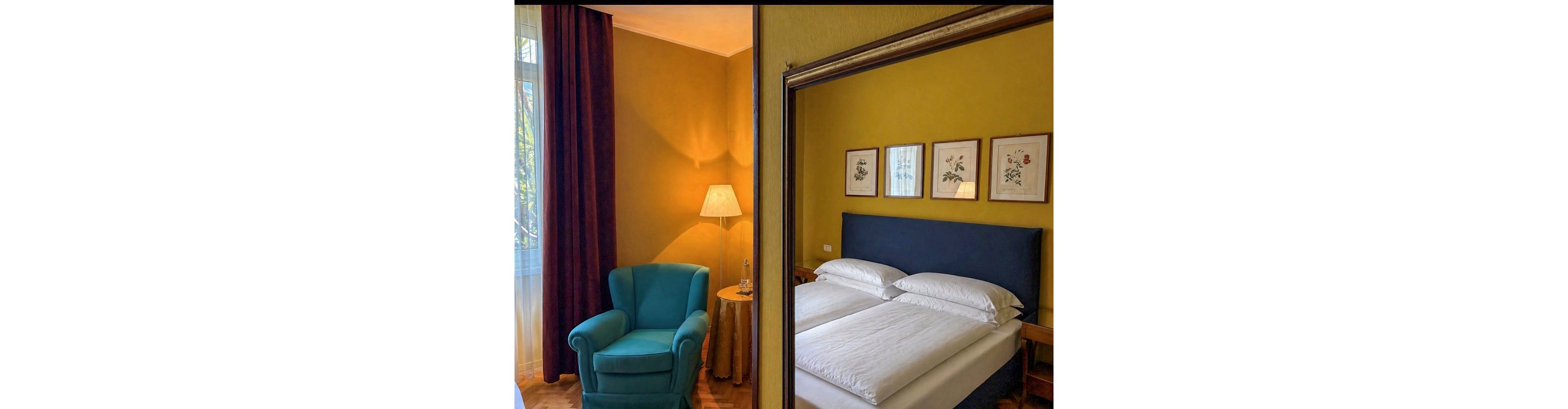 A comfortable hotel room features a teal armchair beside a round table with a lamp, set against mustard yellow walls. The reflection in the mirror shows a double bed with crisp white linens and a navy blue headboard, along with three botanical prints above the bed. Natural light streams in from a window with sheer curtains, creating a warm and inviting atmosphere.