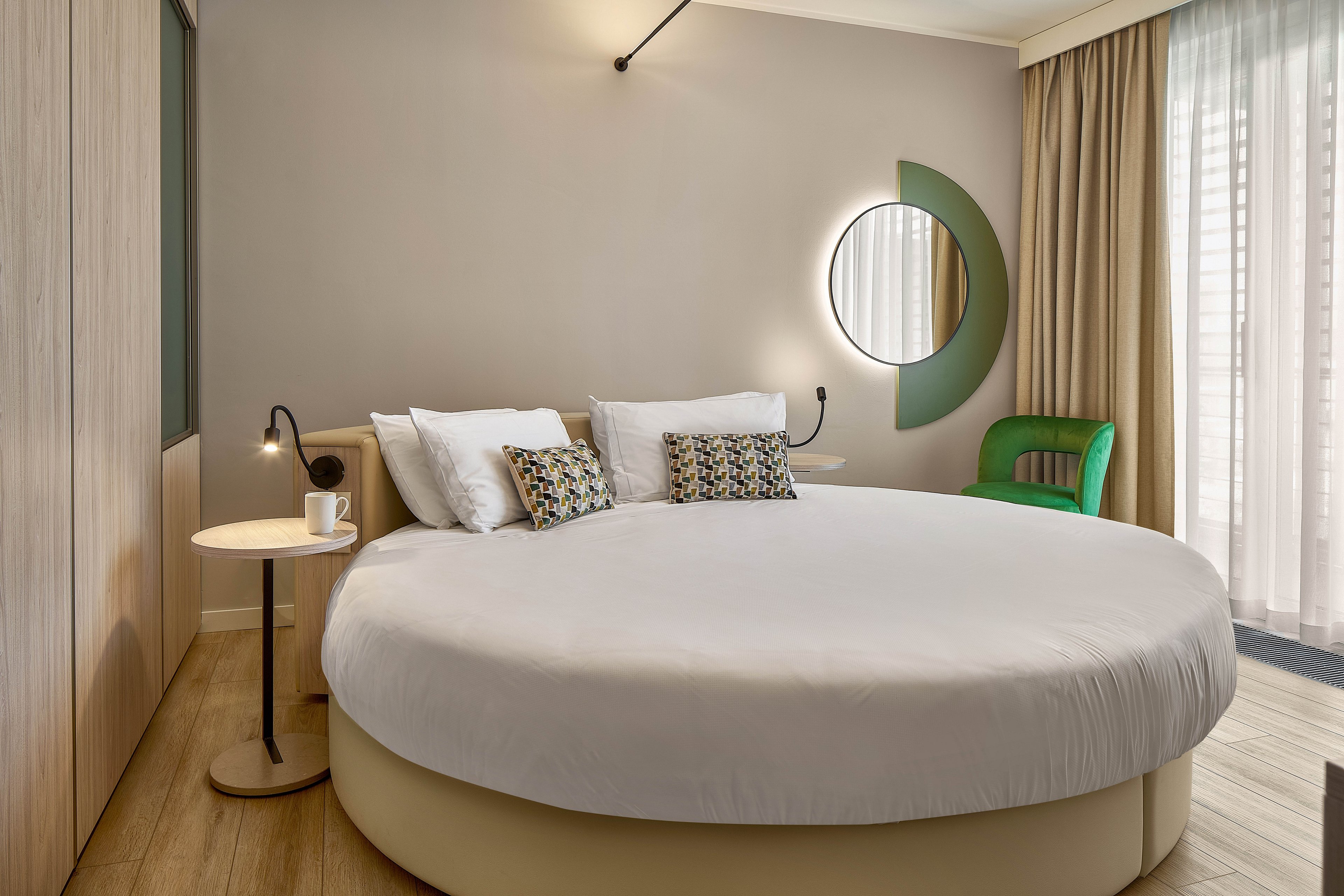 Contemporary hotel bedroom featuring a large round bed with crisp white linens and patterned accent pillows. The space includes a small side table with a cup, wall-mounted reading lights, and a unique circular mirror with a green decorative arc. Floor-to-ceiling curtains filter natural light from the window, complementing the neutral walls and light wood flooring. A green upholstered chair adds a pop of color to the minimalist, stylish interior.